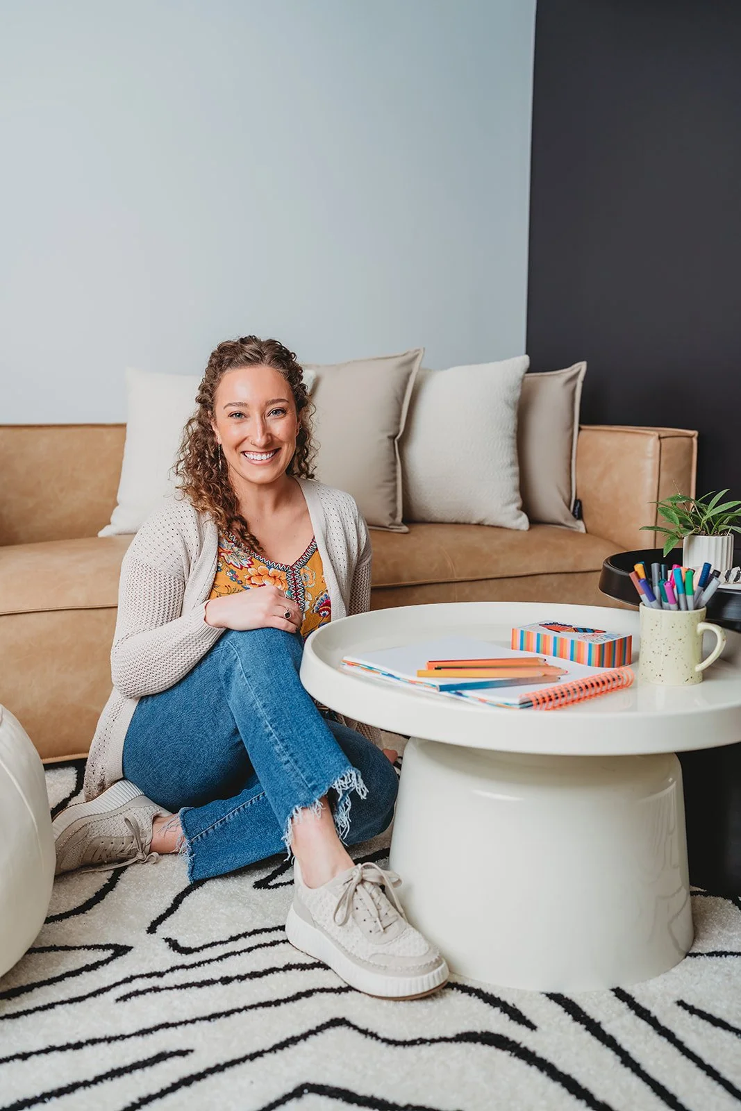 A smiling woman sitting on the floor with her legs crossed in front of a round white coffee table, decorated with school supplies, in a living room with a brown couch and colorful pillows.
