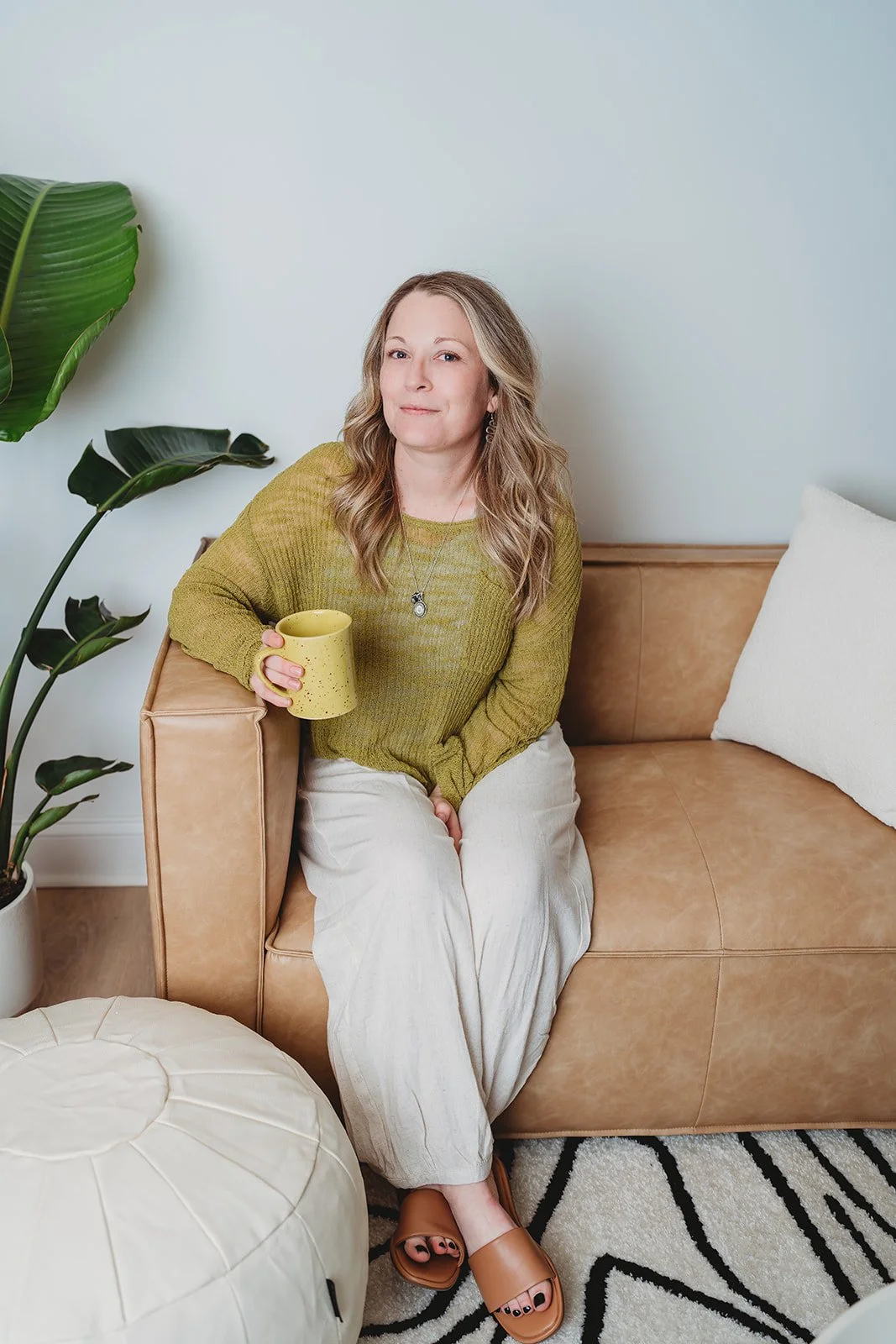 A woman with wavy blonde hair sitting on the floor next to a cream-colored pouf, smiling at the camera. She wears a light gray t-shirt, beige pajamas, and tan slides. There is a tan leather sofa with a white pillow, a large green plant in a white pot, and a white striped rug with black abstract lines in the background.