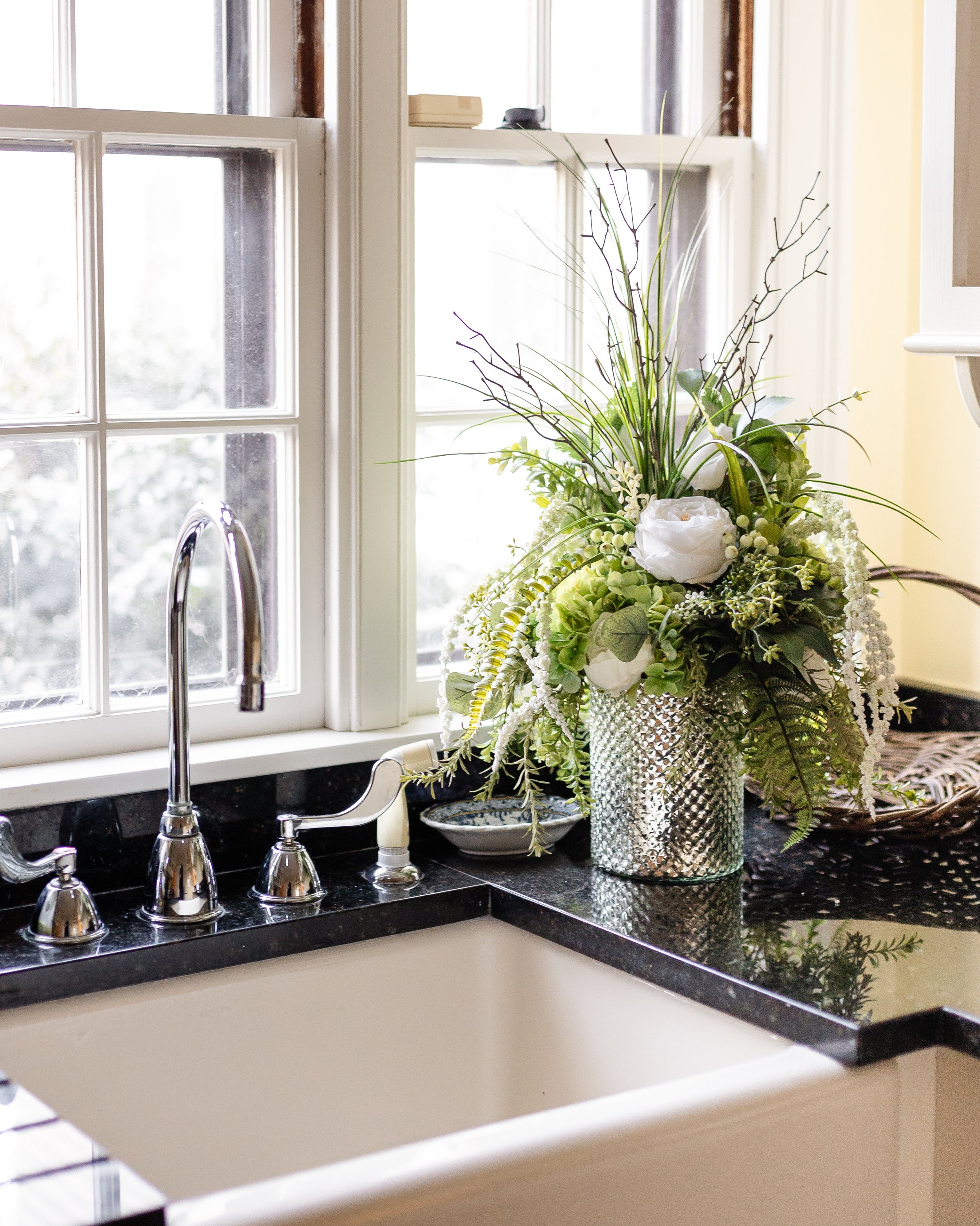 A kitchen window sill with a large floral arrangement in a silver textured vase next to the sink. Designed by Flower Fauxnatic.