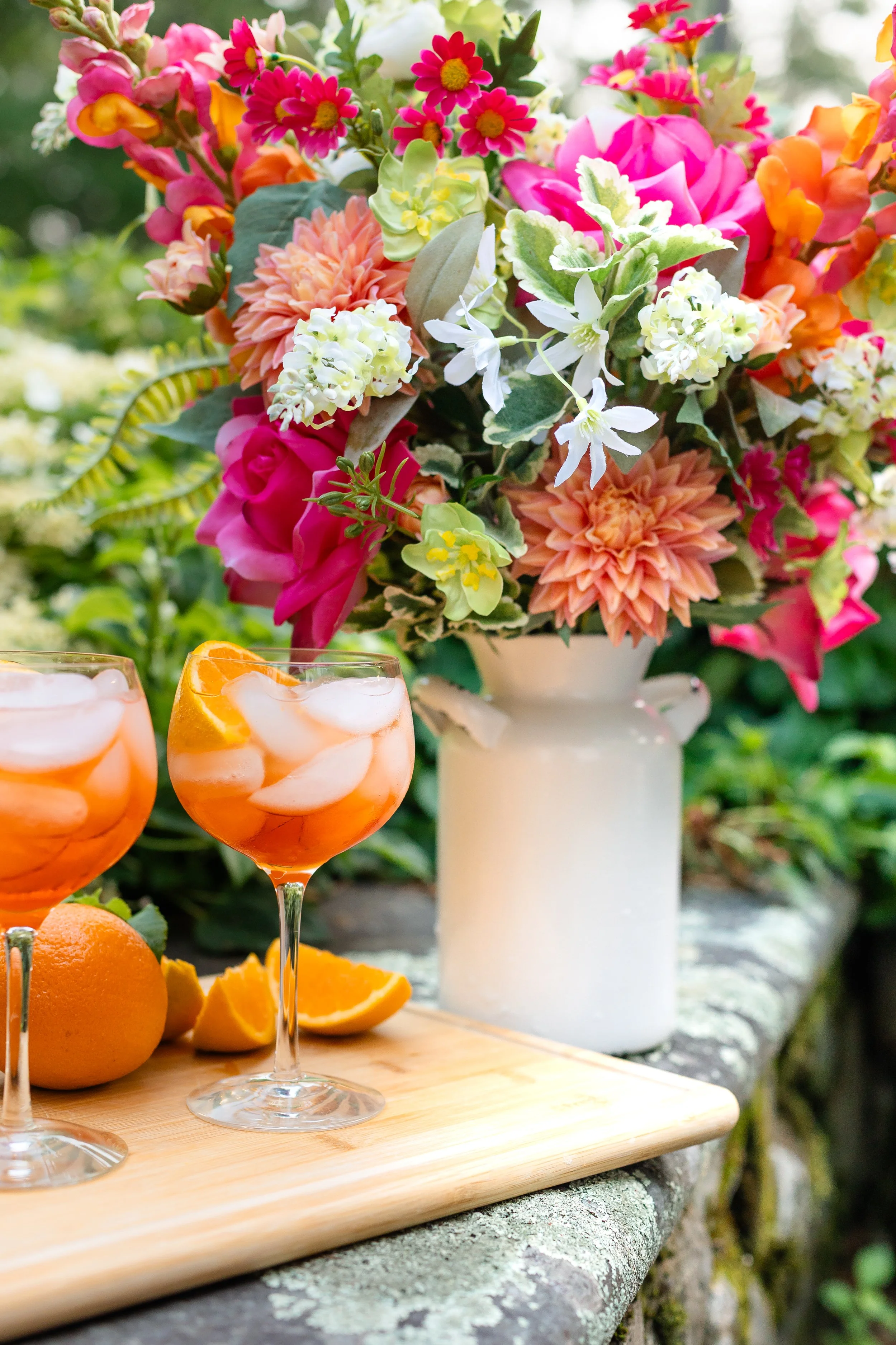 A bouquet of colorful flowers in a white vase beside two glasses of orange-colored drinks with ice and orange slices on a wooden tray, with oranges and slices on the tray. Designed by Flower Fauxnatic.