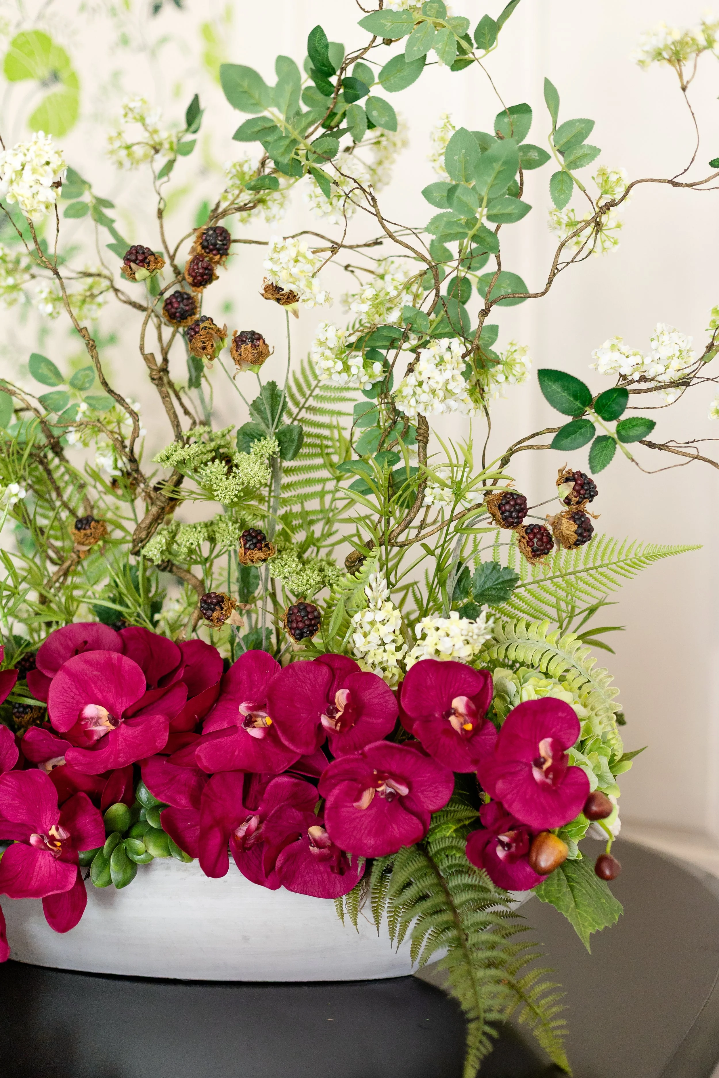 A floral arrangement featuring burgundy orchids, white flowers, blackberries, green ferns, and various green leaves in a white oval vase. Designed by Flower Fauxnatic.