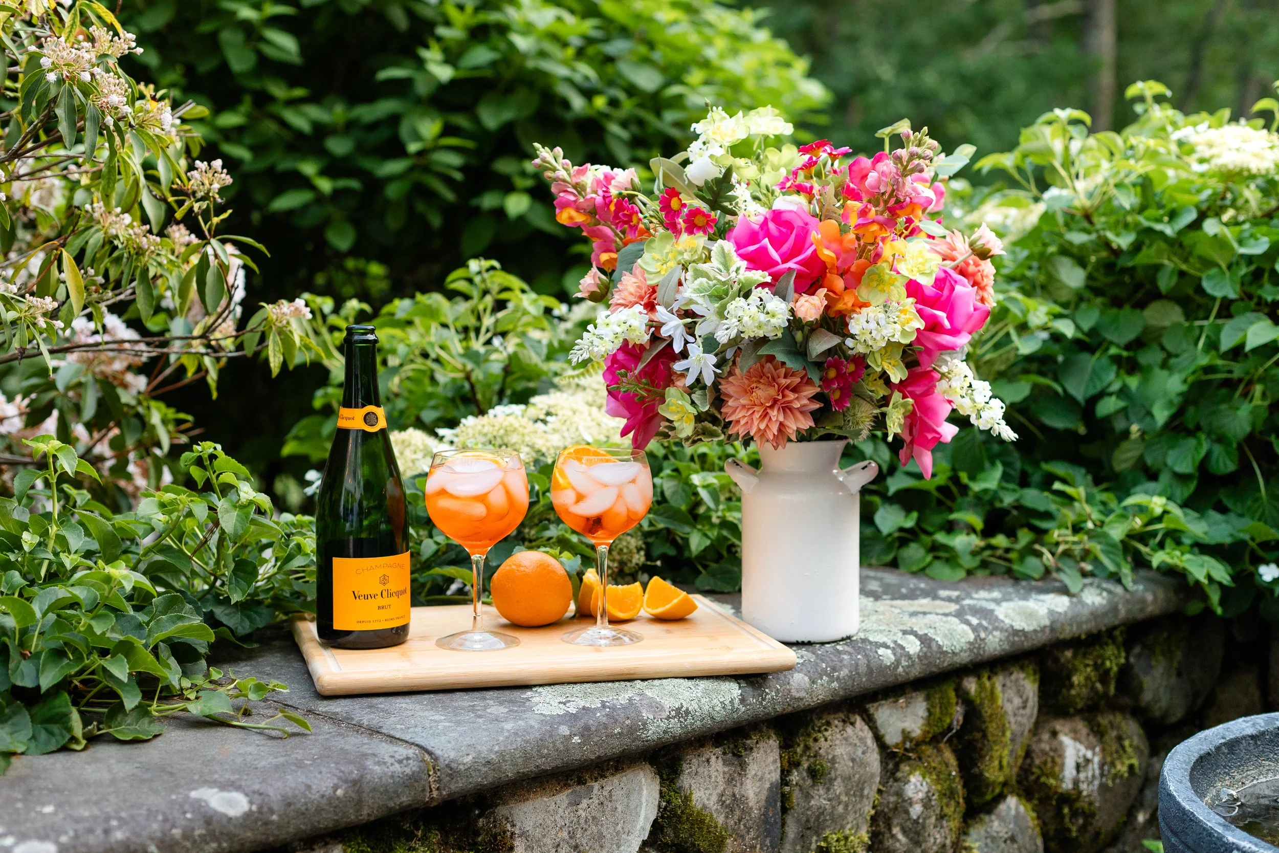 A bottle of Veuve Clicquot champagne, two glasses of orange-colored cocktails with ice and orange slices, a whole orange and orange wedges on a wooden tray, and a white vase filled with pink, yellow, and white flowers. Designed by Flower Fauxnatic.