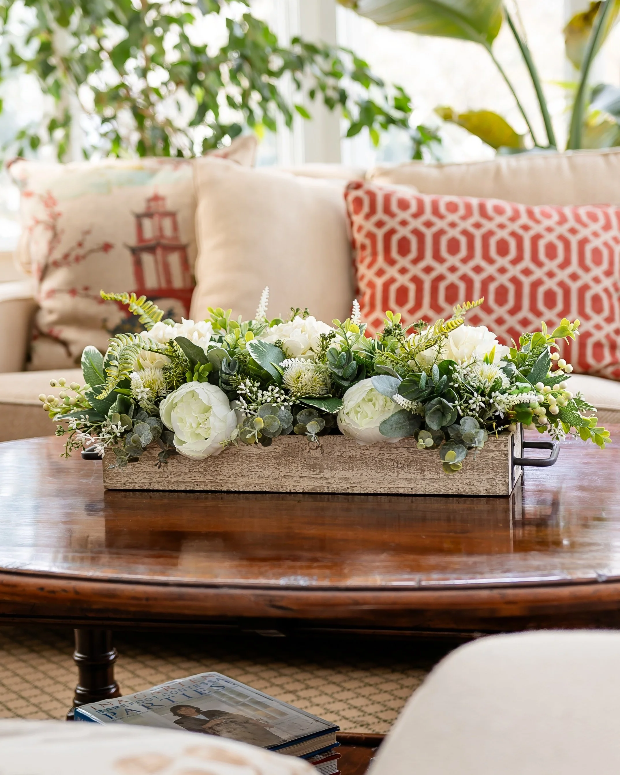 A decorative floral arrangement on a wooden coffee table in a cozy living room with beige and patterned pillows and green houseplants in the background. Designed by Flower Fauxnatic.