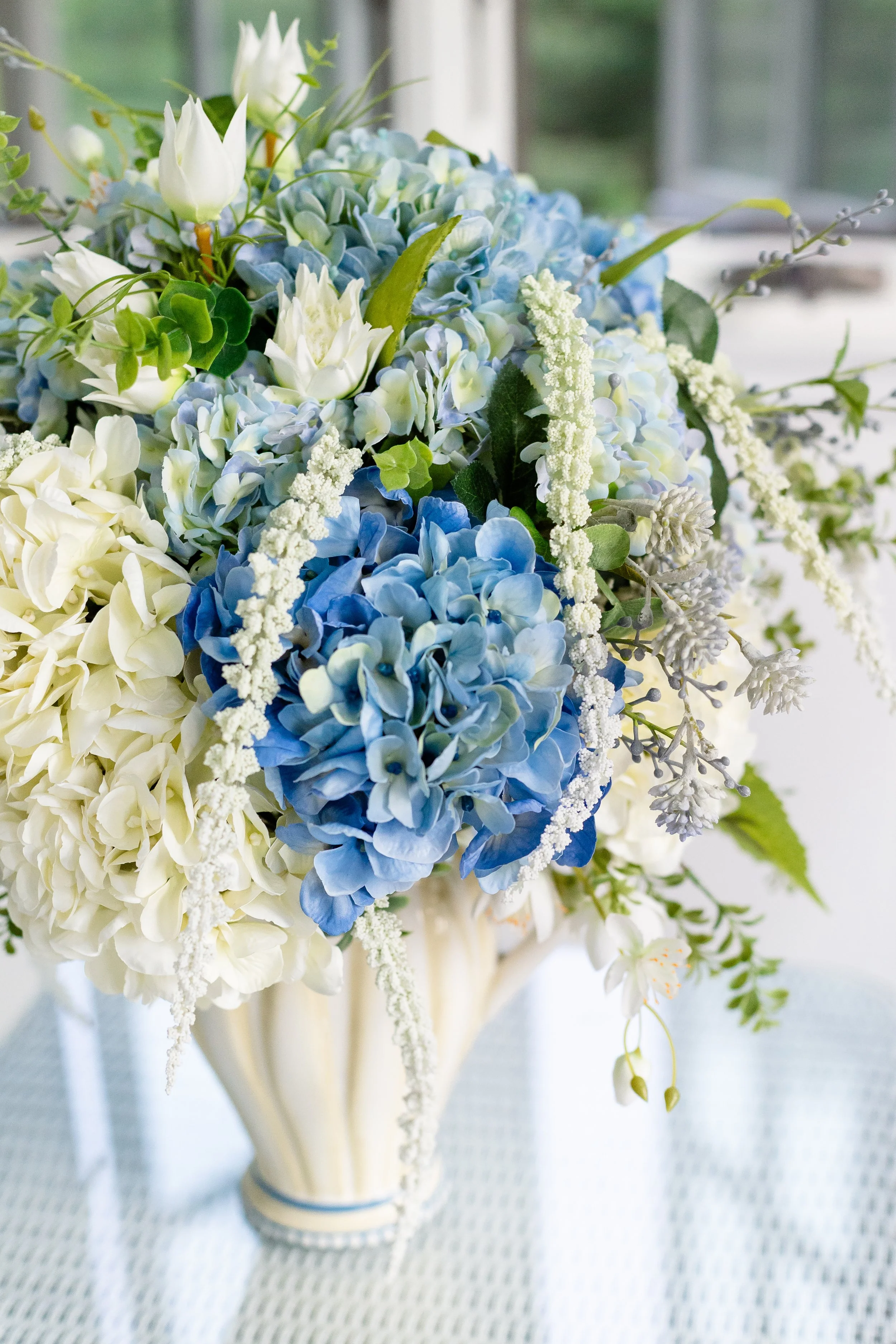 A floral arrangement with white and blue flowers in a cream-colored vase. Designed by Flower Fauxnatic.