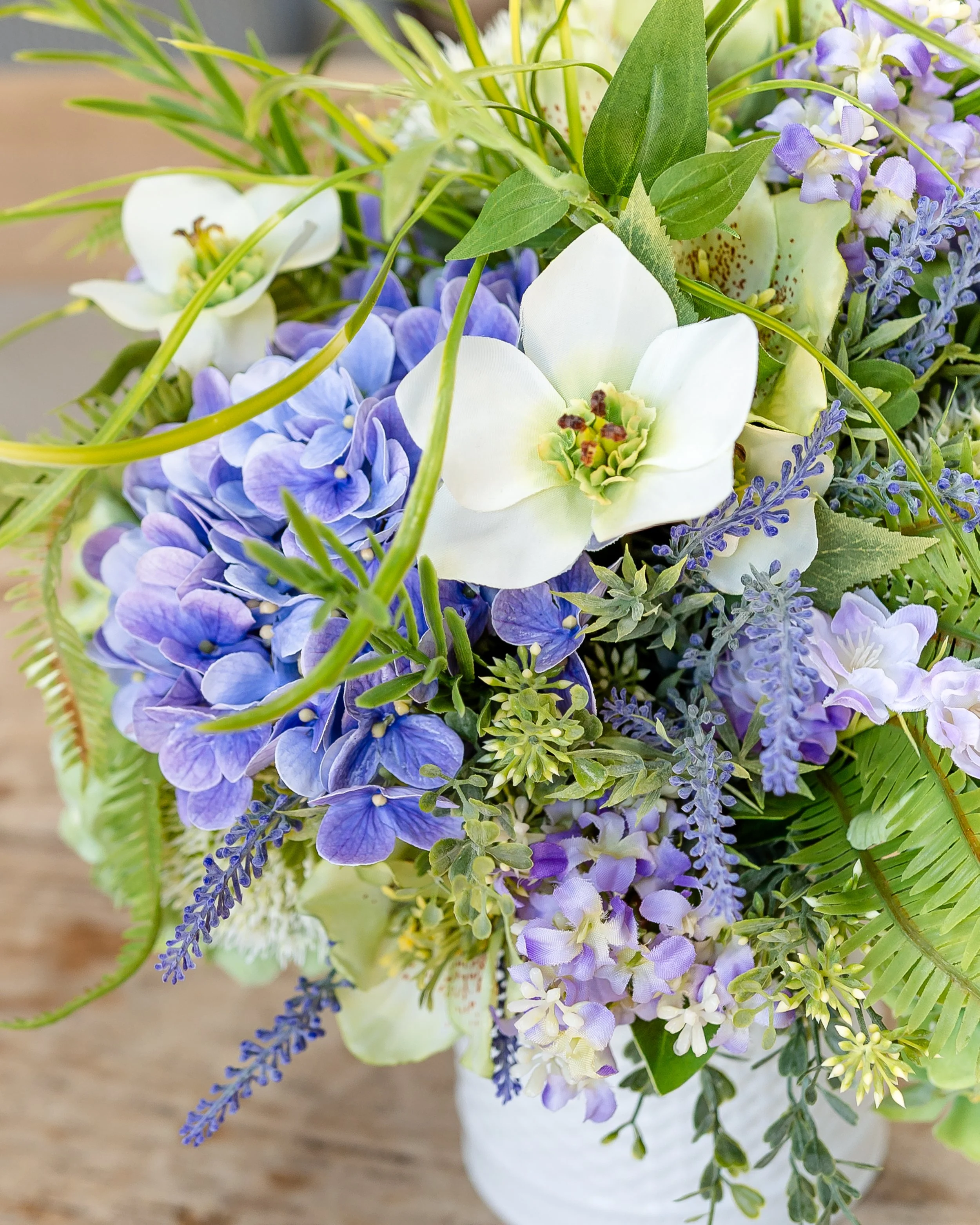 Close-up of a bouquet of mixed purple, white, and green flowers with greenery, in a white vase on a wooden surface. Designed by Flower Fauxnatic.
