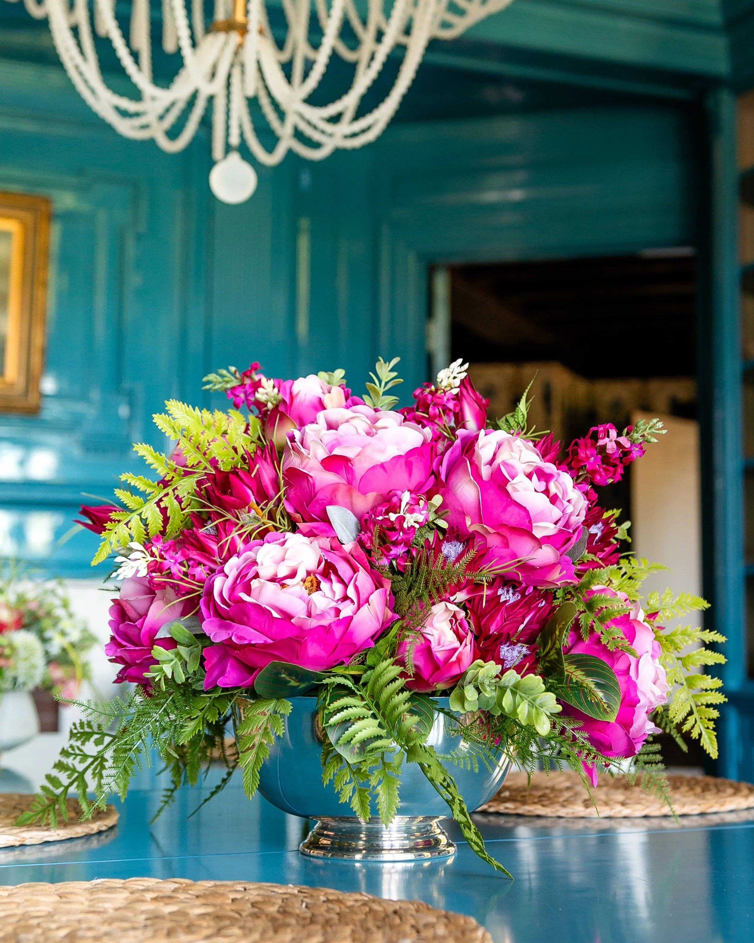 A vibrant bouquet of pink and white peonies and other flowers in a silver bowl, placed on a blue table with woven placemats, set against a teal-painted wooden interior with a white chandelier overhead. Designed by Flower Fauxnatic.