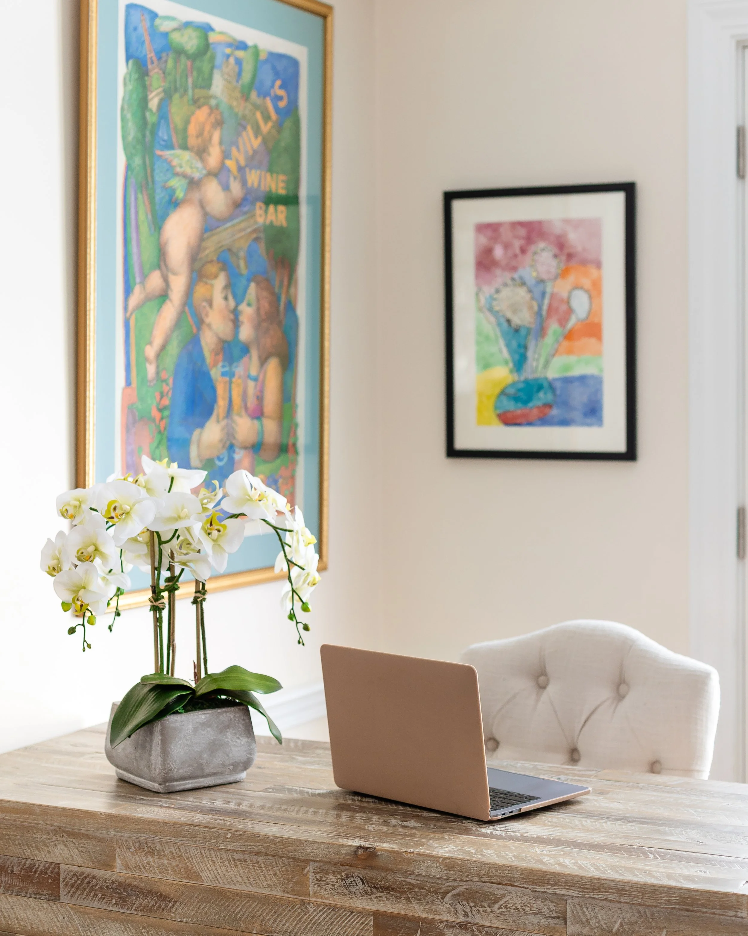 A desk with a potted white orchid, a pink laptop, a beige chair, and framed colorful artwork on a light-colored wall. Designed by Flower Fauxnatic.
