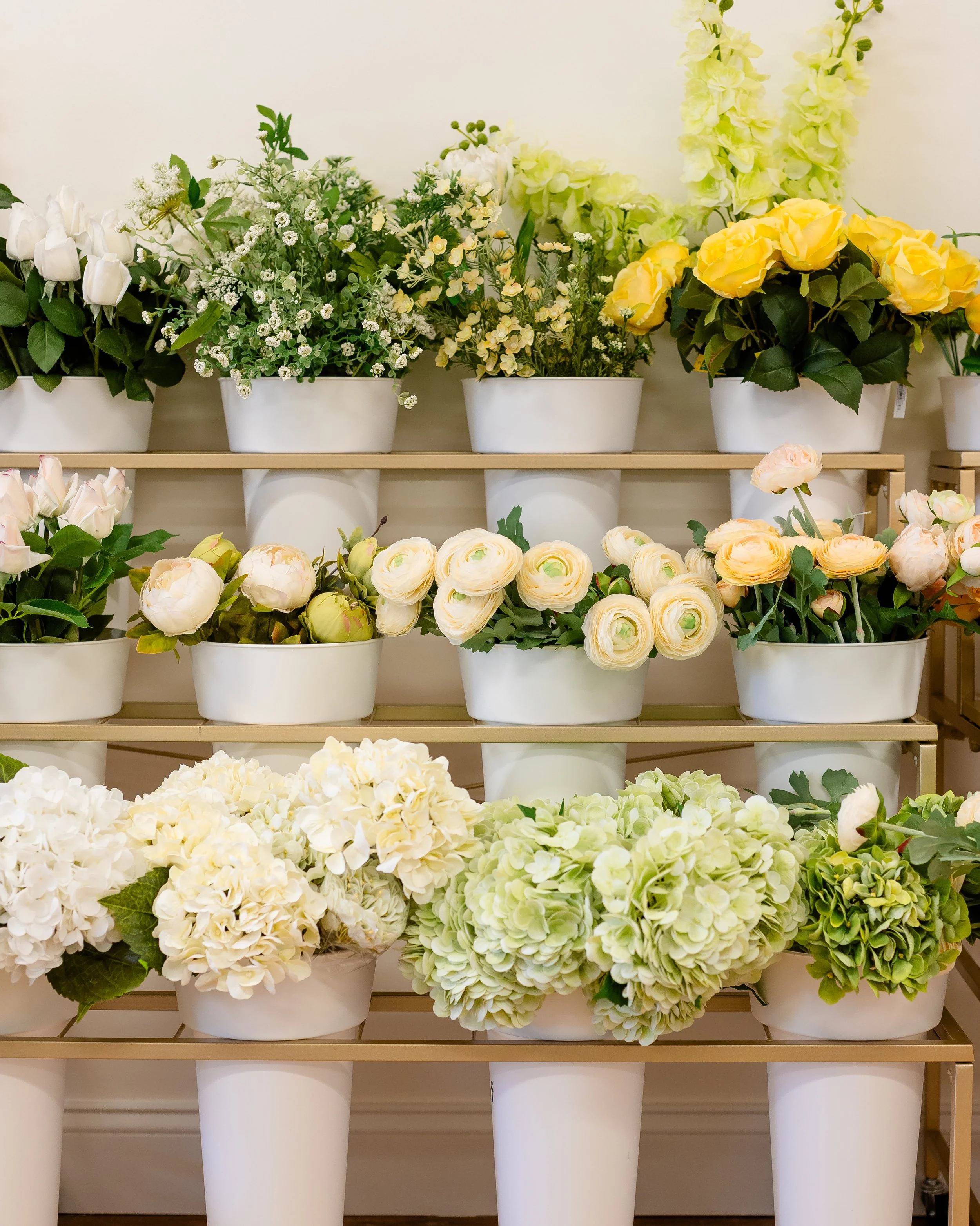 Display of white flower pots filled with white and light-colored roses, hydrangeas, and other flowers on wooden shelves against a white wall.