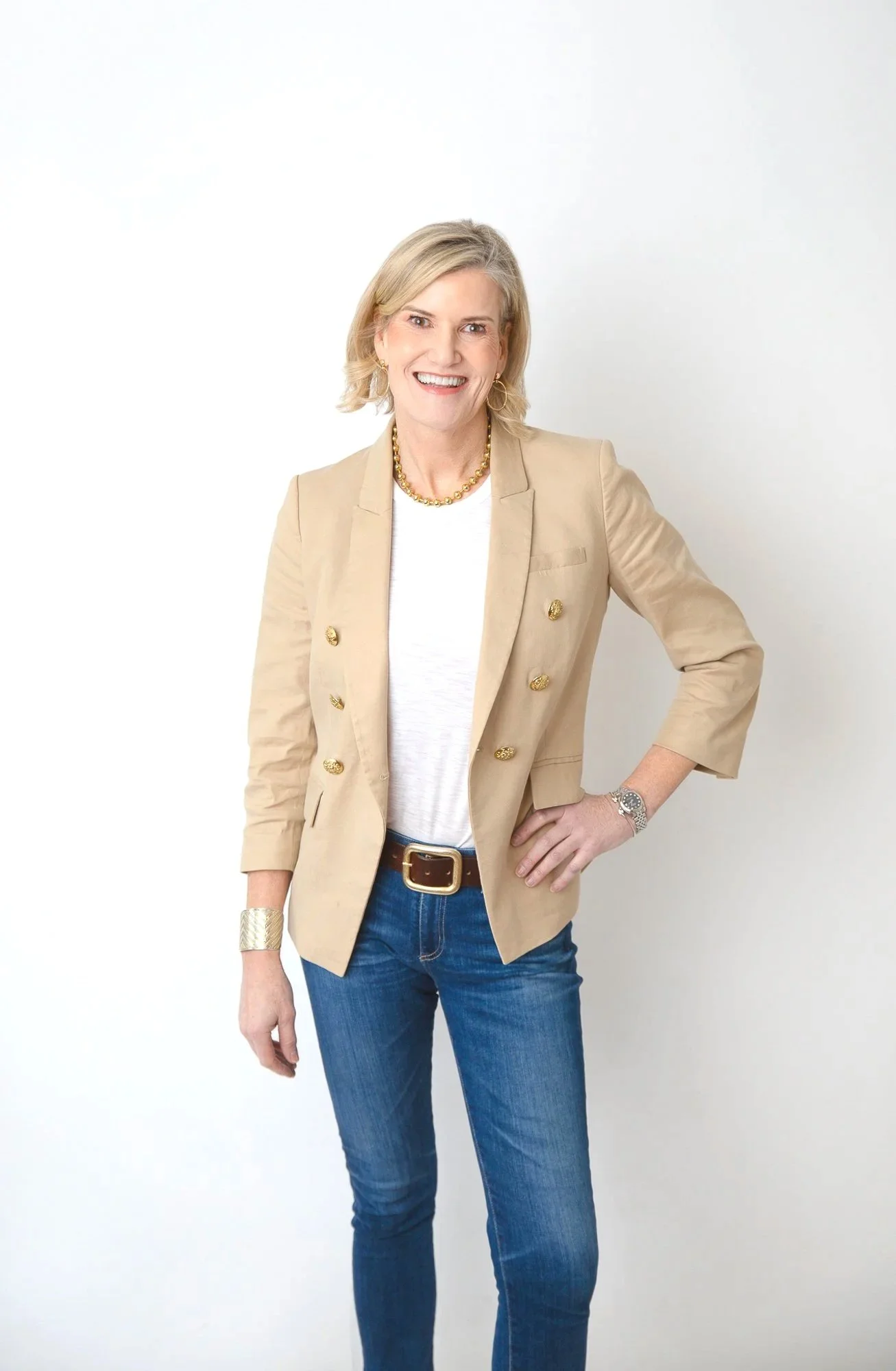 Alex, founder of Flower Fauxnatic, wearing a beige blazer, white top, blue jeans, and jewelry, standing against a plain white background and smiling at the camera.