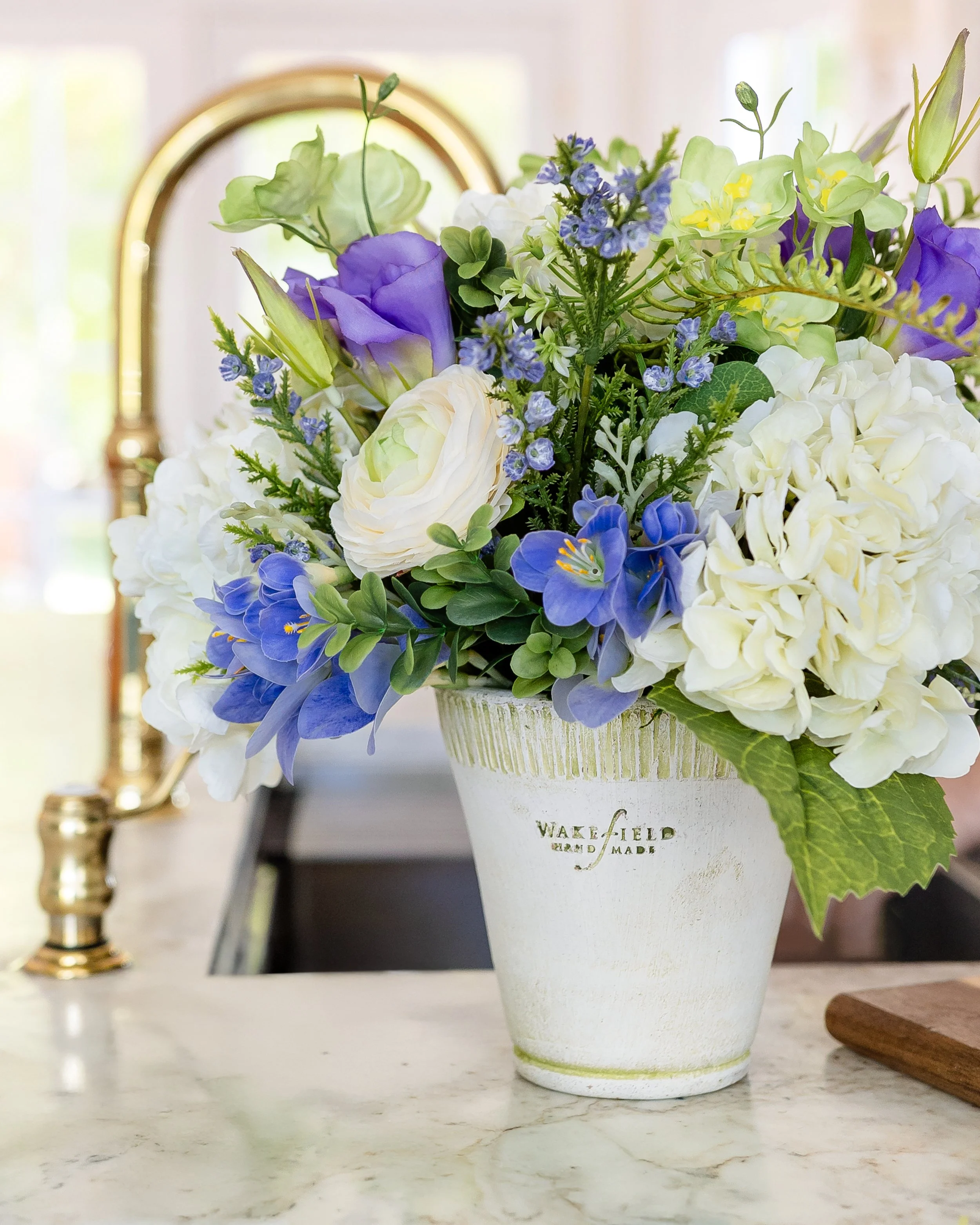 A floral arrangement with white, blue, and purple flowers in a white pot on a kitchen island. Designed by Flower Fauxnatic.