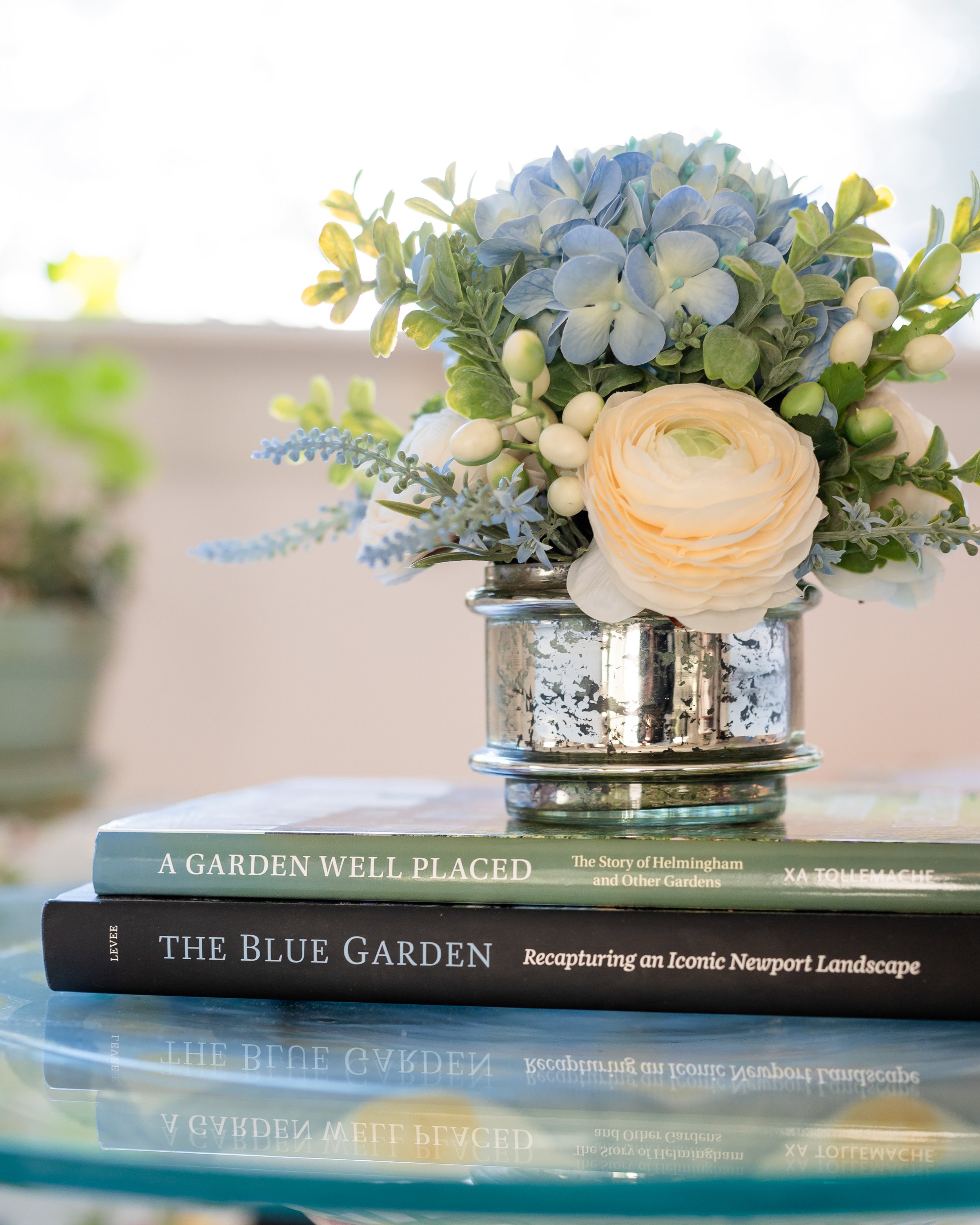 A bouquet of blue, white, and green flowers in a reflective silver vase, placed on top of two gardening books about gardens and landscapes. Designed by Flower Fauxnatic.