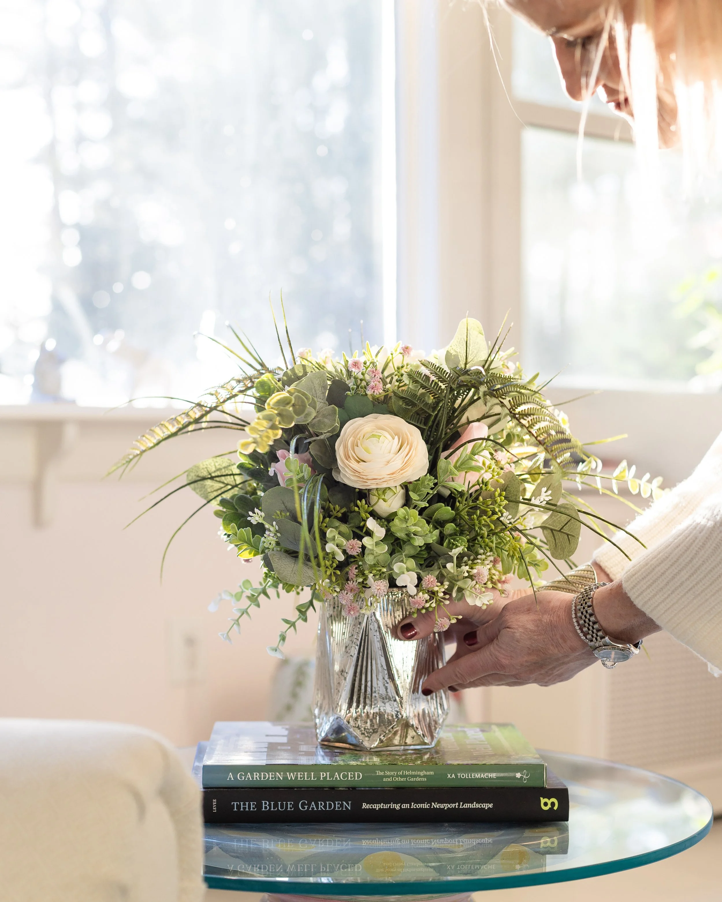 A woman arranging a bouquet of flowers in a glass vase on a table, with books underneath, near a window. Designed by Flower Fauxnatic.