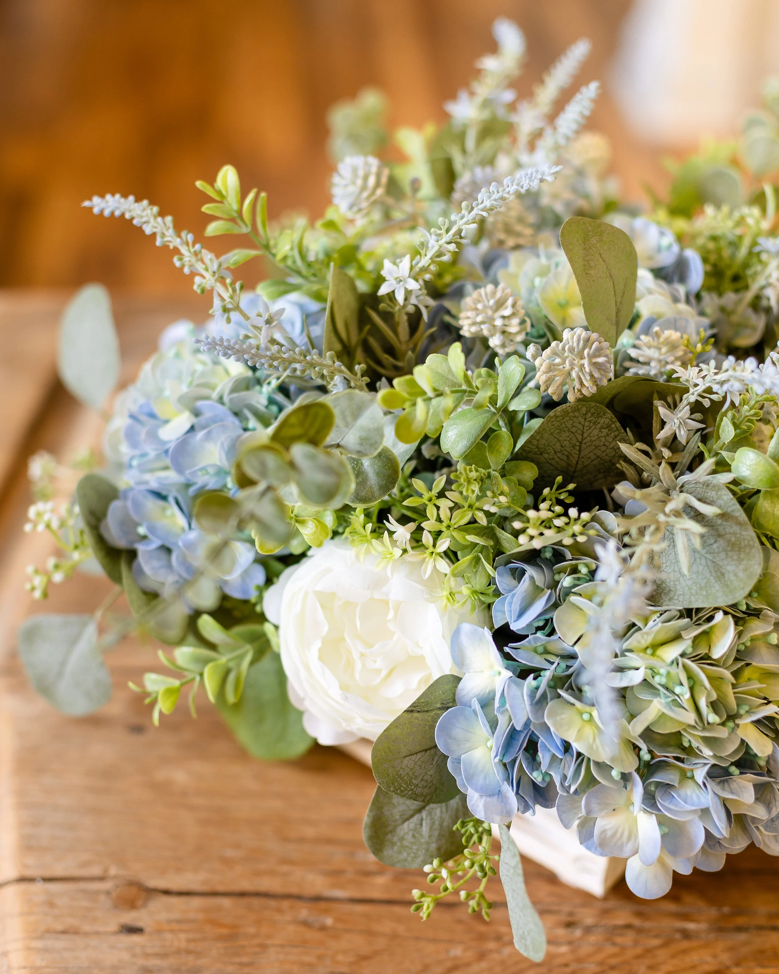 A close-up of a bouquet with white and light blue flowers and green leaves on a wooden surface. Designed by Flower Fauxnatic.