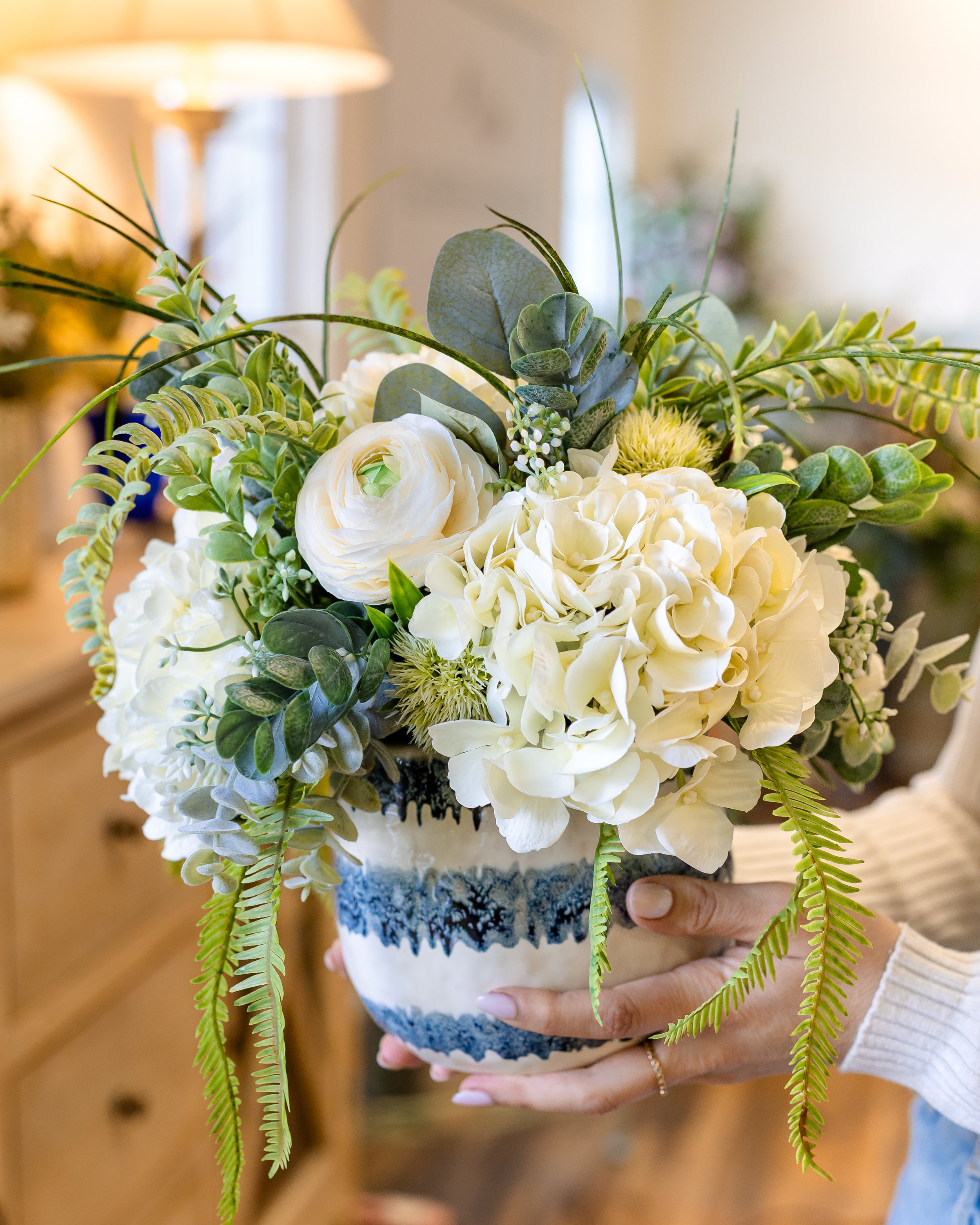 Person holding a white and blue patterned ceramic vase filled with white flowers and green foliage. Designed by Flower Fauxnatic.