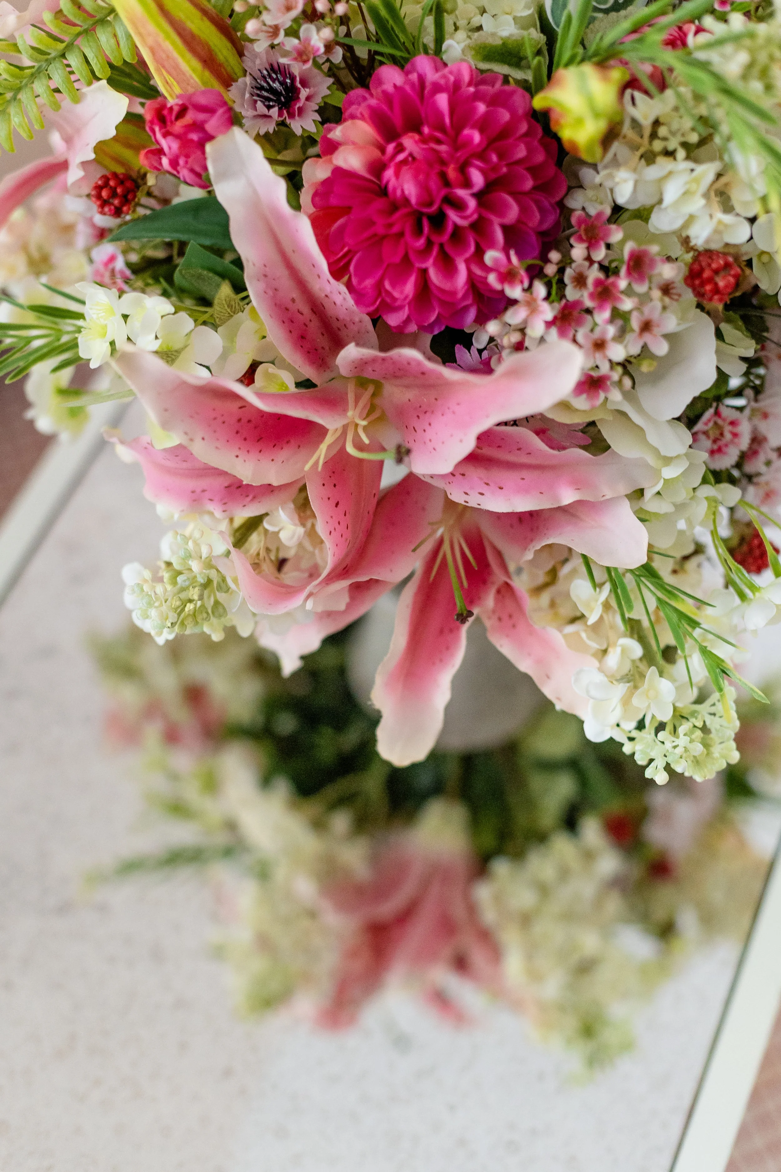 Close-up of a colorful bouquet of flowers including pink lilies, red dahlias, small white flowers, and green foliage. Designed by Flower Fauxnatic.