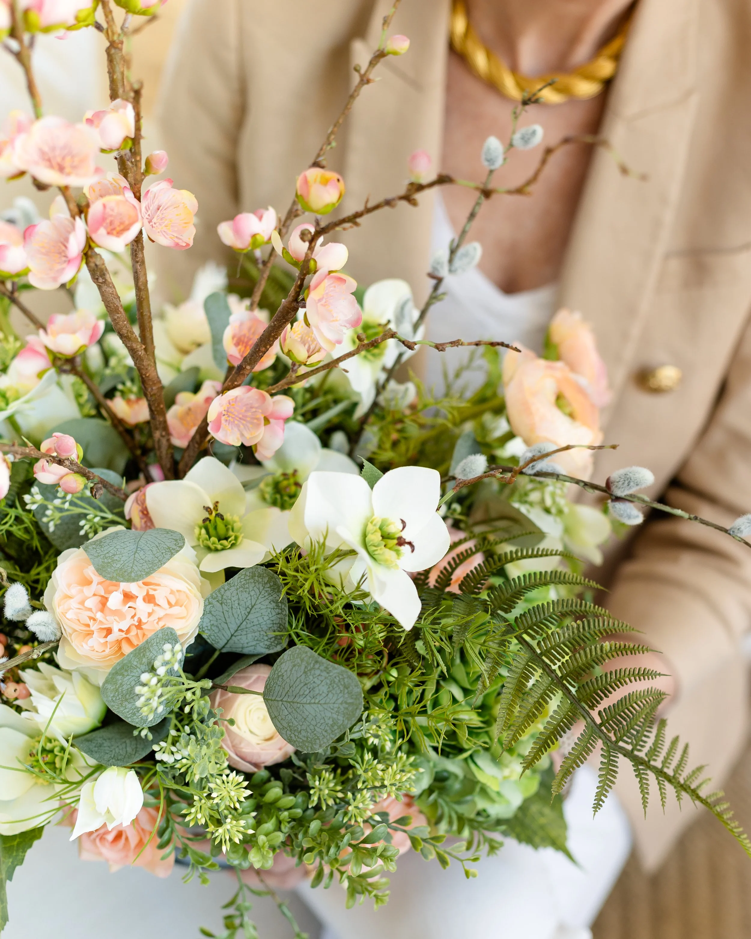 Close-up of a floral arrangement featuring pink cherry blossoms, white dogwood flowers, peach roses, green ferns, and eucalyptus leaves, with a person in a beige blazer holding it. Designed by Flower Fauxnatic.