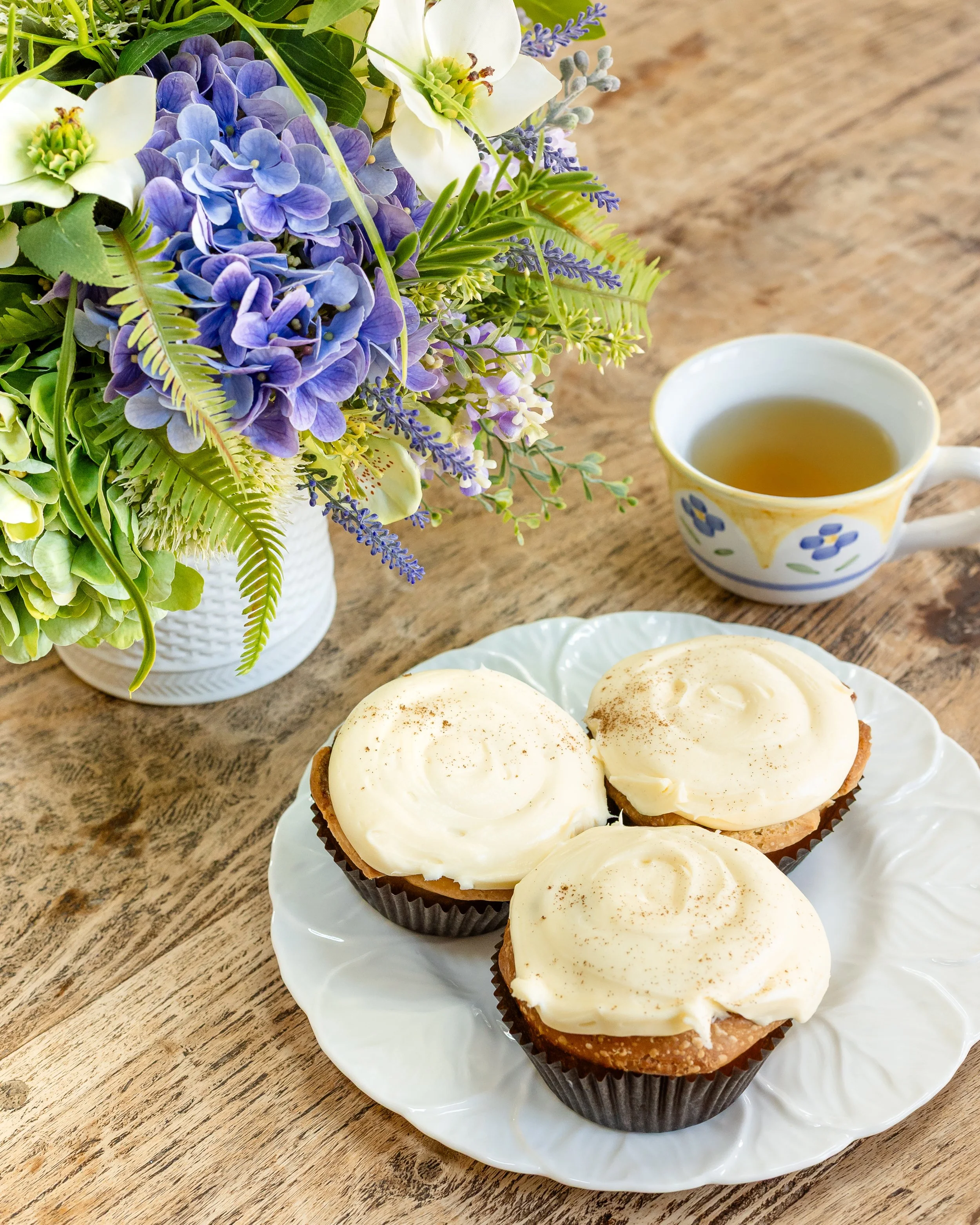A white plate with three frosted cupcakes on a wooden table, a cup of tea, and a vase of purple, white, and green flowers. Designed by Flower Fauxnatic.