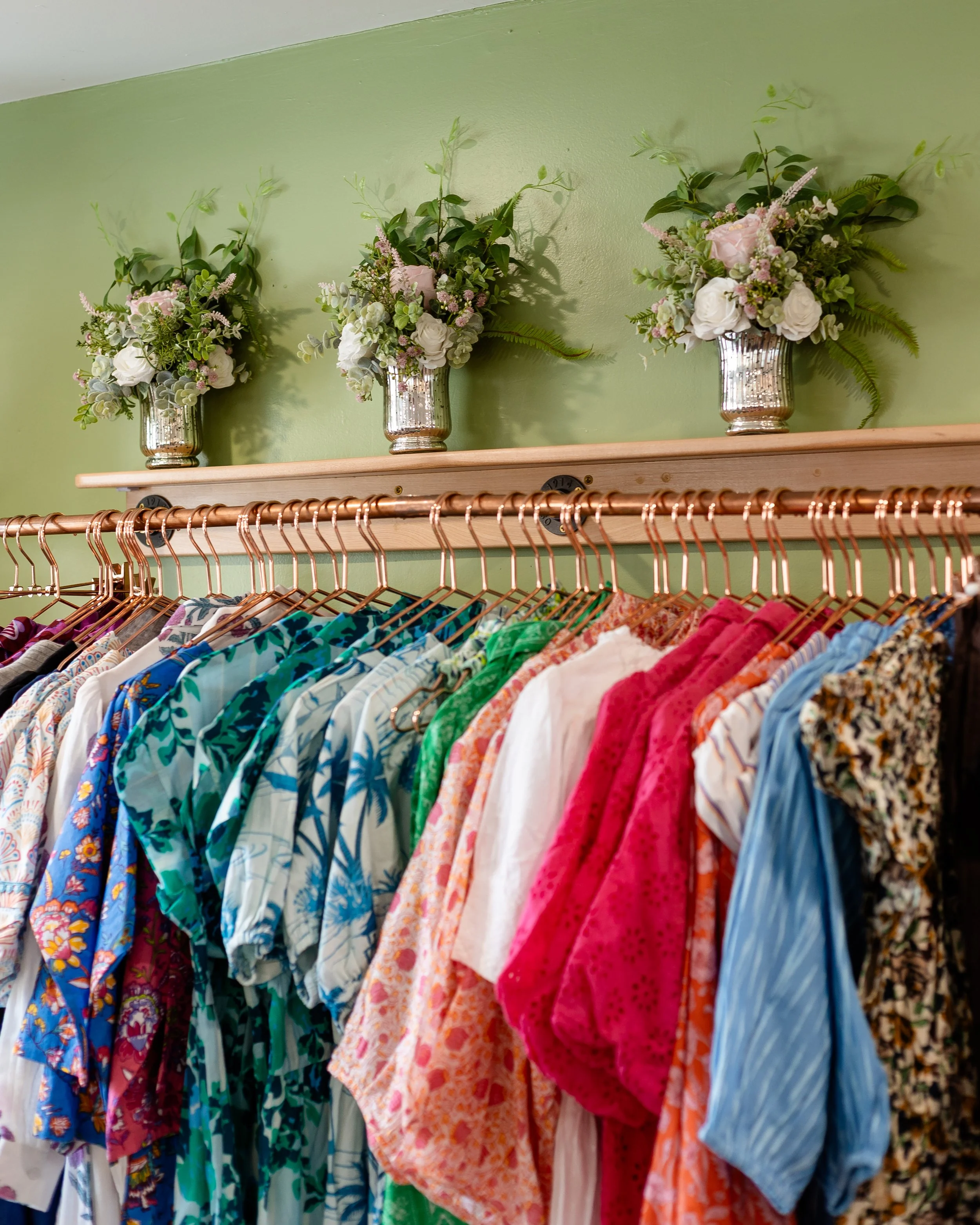 Colorful clothing hanging on a copper rack with three floral arrangements in silver vases on a green wall shelf above. Designed by Flower Fauxnatic.