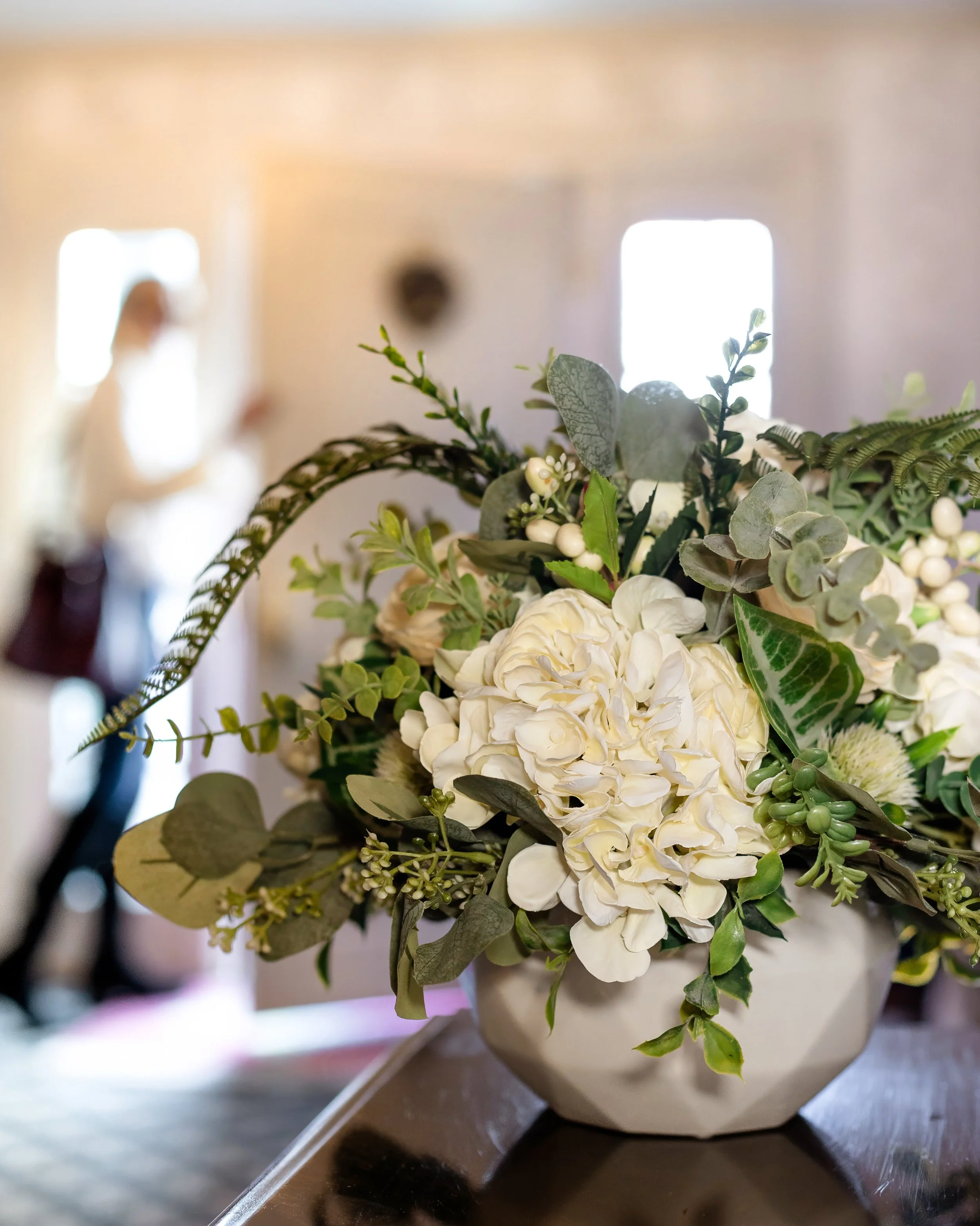 A white ceramic vase filled with white hydrangeas, green leaves, and foliage on a wooden surface, with a blurred person in the background. Designed by Flower Fauxnatic.