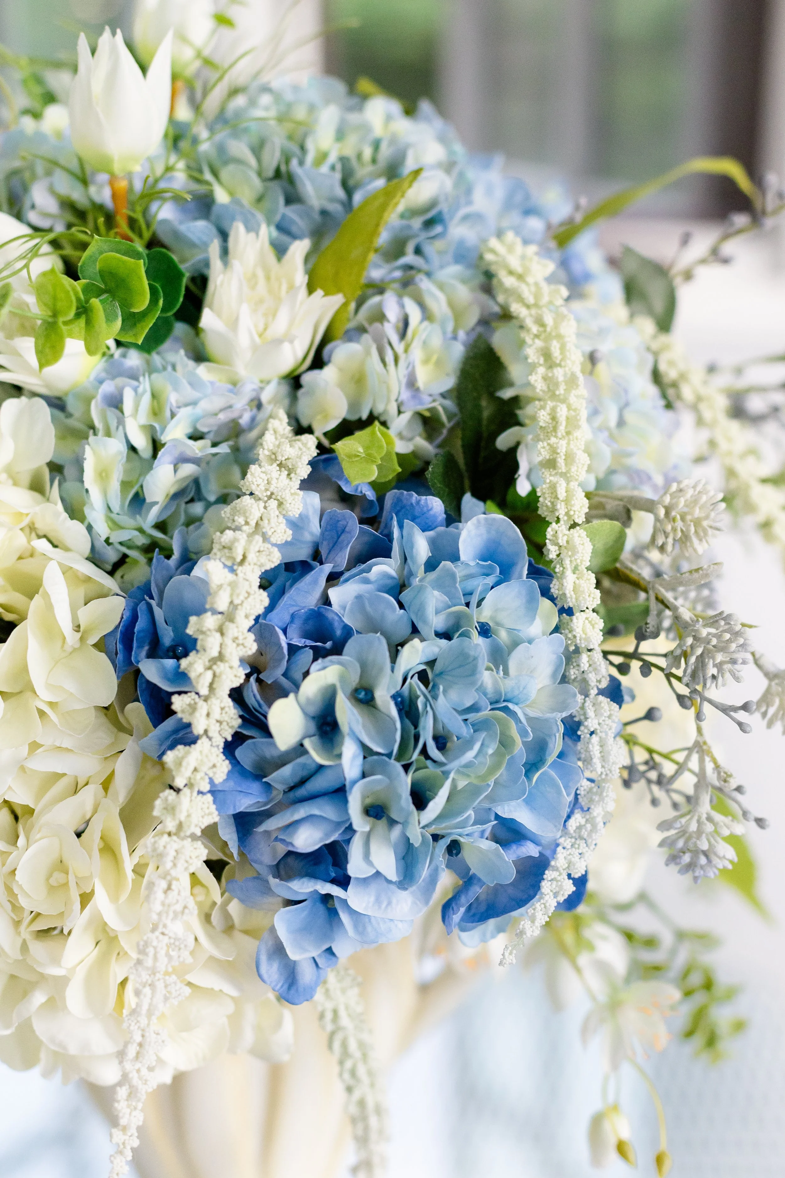 Close-up of a bouquet with blue and white hydrangeas, white tulips, and other white flowers with green leaves. Designed by Flower Fauxnatic.