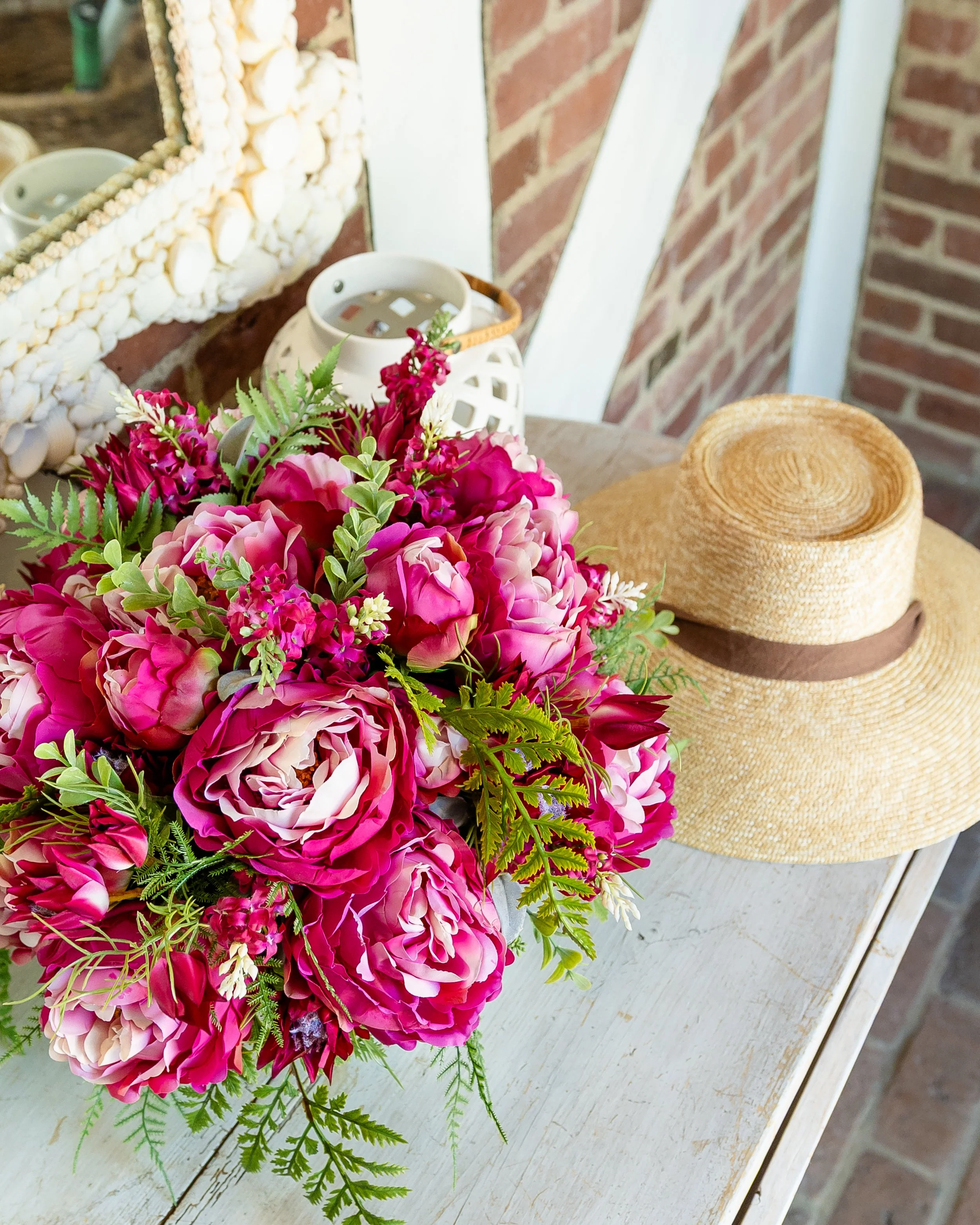 A bouquet of pink and purple flowers on a white wooden table, with a straw hat and a white candle holder nearby, against a brick wall background. Designed by Flower Fauxnatic.