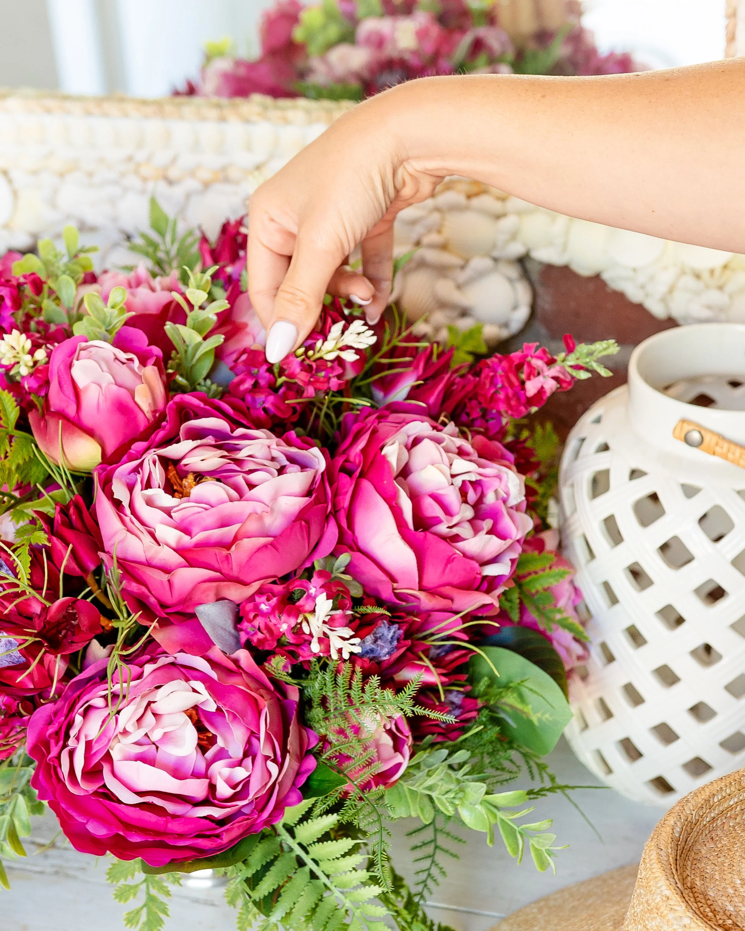 A person's hand arranging a pink and purple flower bouquet with various green foliage, next to a white wicker lantern and a woven straw hat. Designed by Flower Fauxnatic.