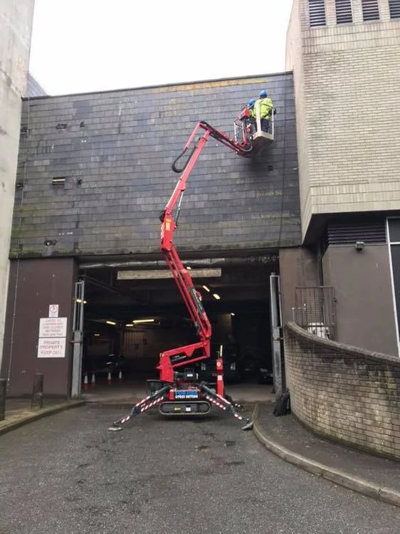 A worker in a bucket lift inspecting or working on the upper part of a building's exterior wall, which is covered with dark shingles. The worker is wearing a yellow safety vest and a helmet. The lift is positioned in front of a parking garage entranc