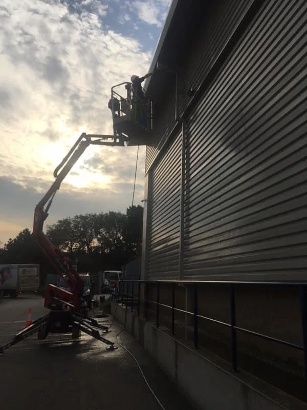 A worker in a cherry picker is cleaning or inspecting the roof of a large industrial building with metallic siding during sunset.