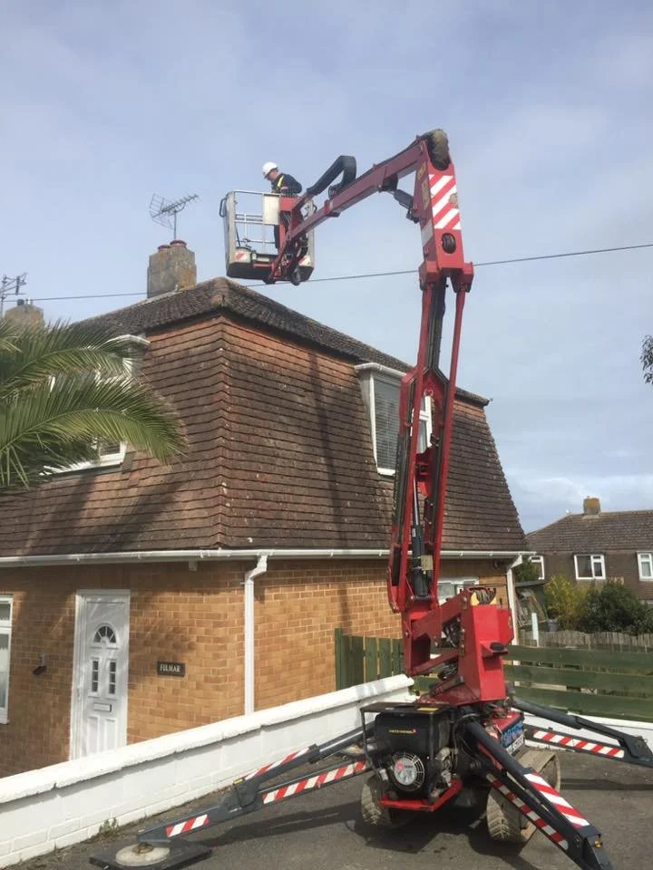 A worker in a safety helmet is using a cherry picker to work on the roof of a brick house.