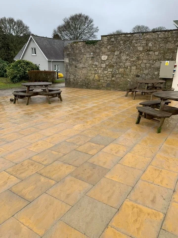 An outdoor patio with yellow-brown stone tiles and four wooden picnic tables. There is a high stone wall on one side and trees and a house in the background.