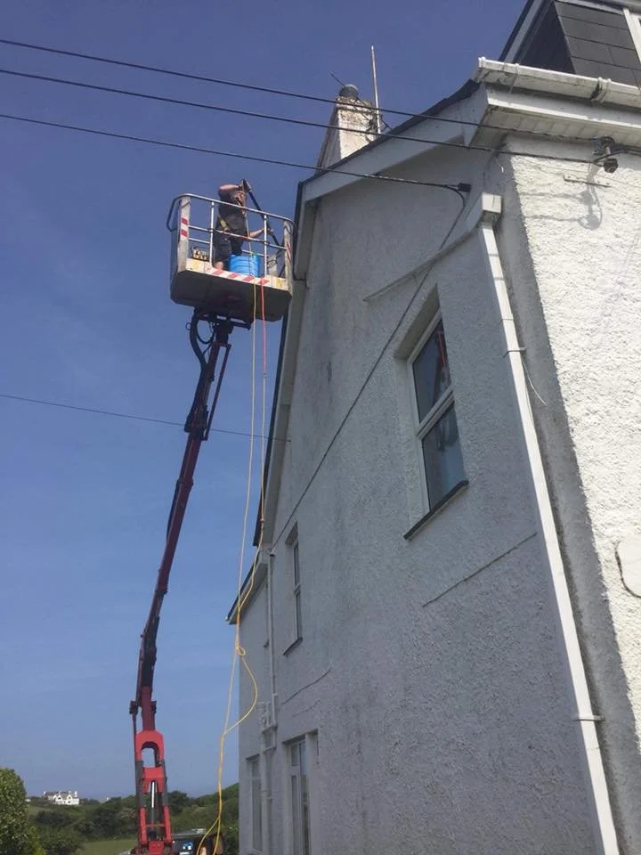 A worker on an elevated lift inspecting or repairing electrical wires on the side of a white house against a clear blue sky.