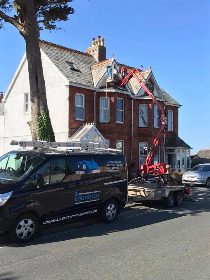 Workers using a cherry picker to clean or repair the roof of a large, red-brick house with a white upper facade. A black service van with company information is parked nearby.