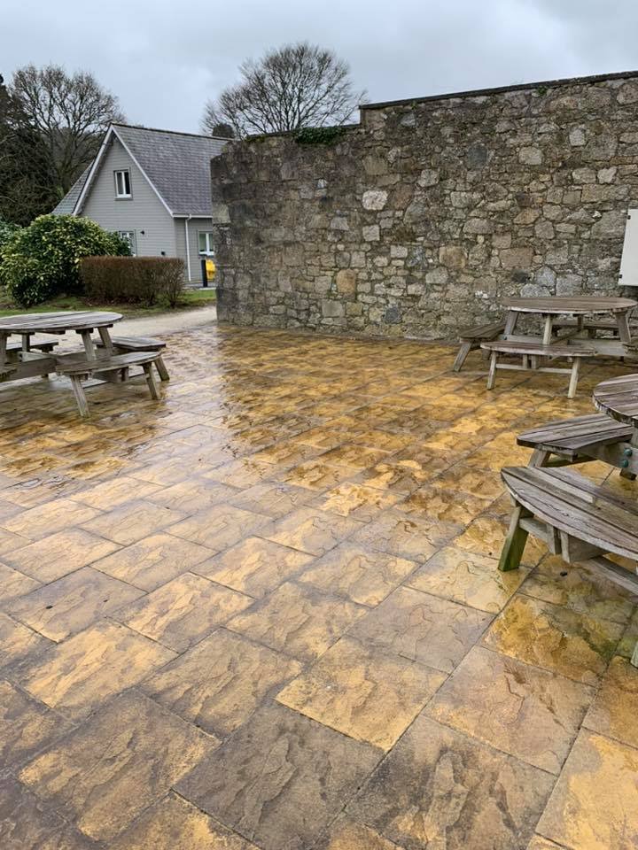 Wet outdoor patio with stone tiles, wooden picnic tables, stone wall, neighboring house, trees, and cloudy sky.