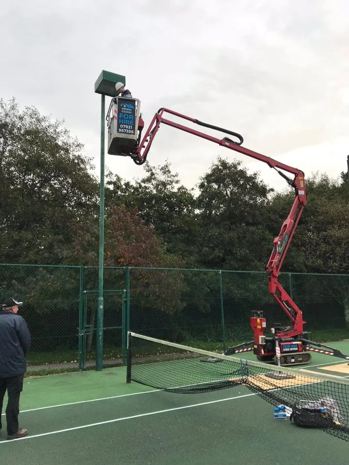 Worker on a cherry picker inspecting a streetlight on a tennis court surrounded by a chain-link fence.