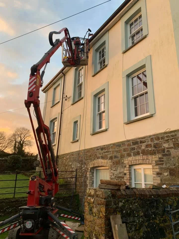 A cherry picker lift extended to windows of a three-story house with a stone foundation. The house has cream-colored walls with green shutters, and there are trees and a sunset sky in the background.