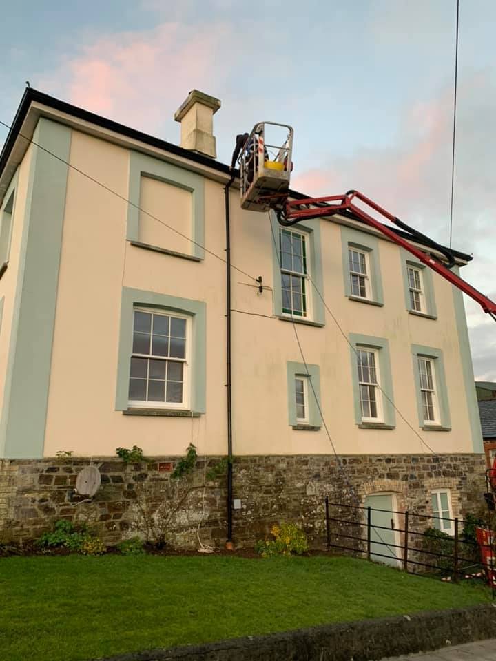 Worker using a cherry picker to paint the upper part of a multi-story house with light-colored walls and green window trims, against a sunset sky.