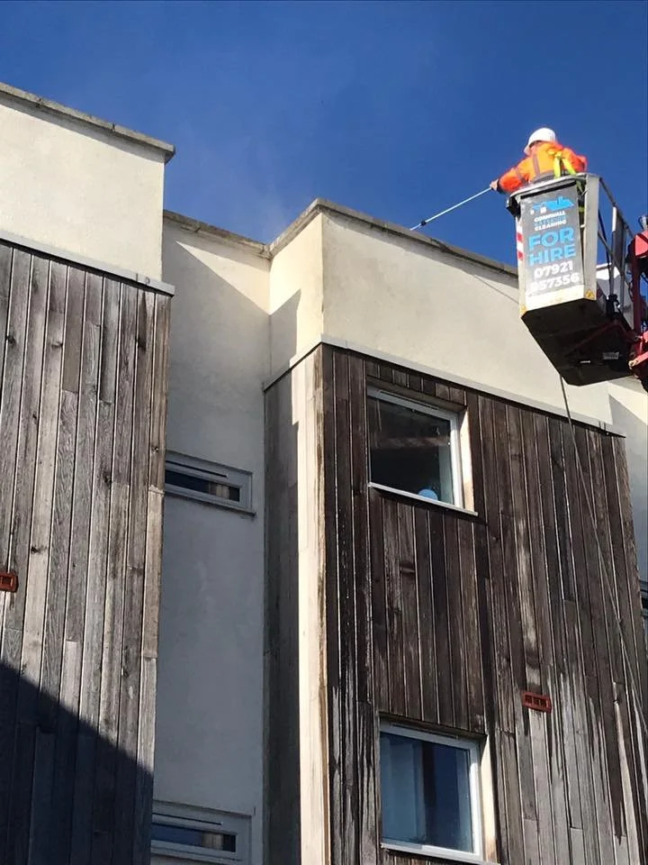 A worker in safety gear on a cherry picker applying weatherproofing or paint to the top edge of a modern multi-story building with a mix of white and dark wood exterior panels under a blue sky.