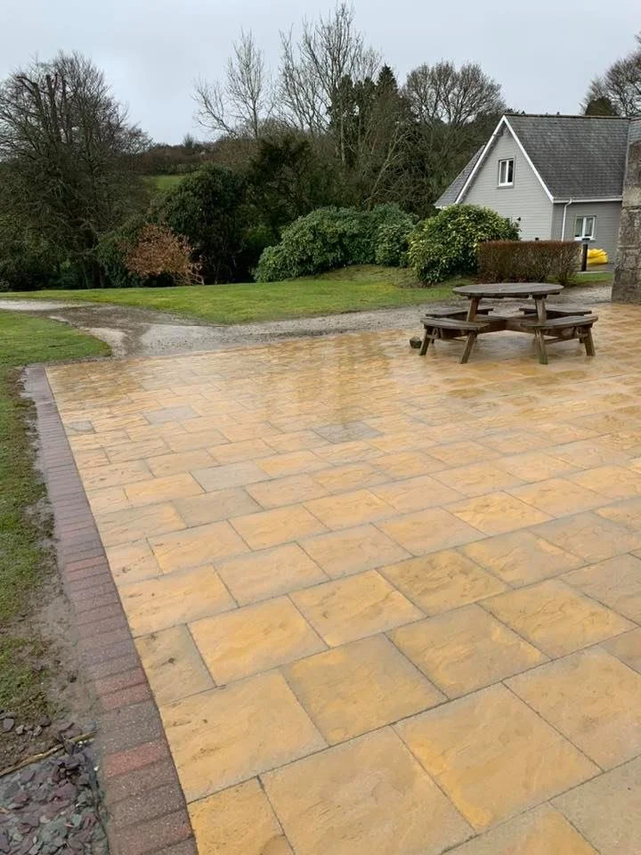 A wet stone patio with a wooden picnic table, background of grass, bushes, and a house with trees and a cloudy sky.