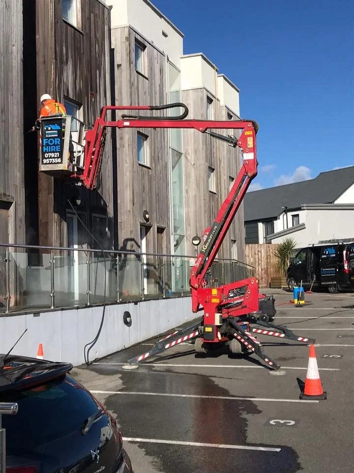 A construction worker in an orange safety vest and white helmet is elevated in a cherry picker working on the exterior of a multi-story building. The building has a wooden facade and large windows. There are orange safety cones around the cherry pick