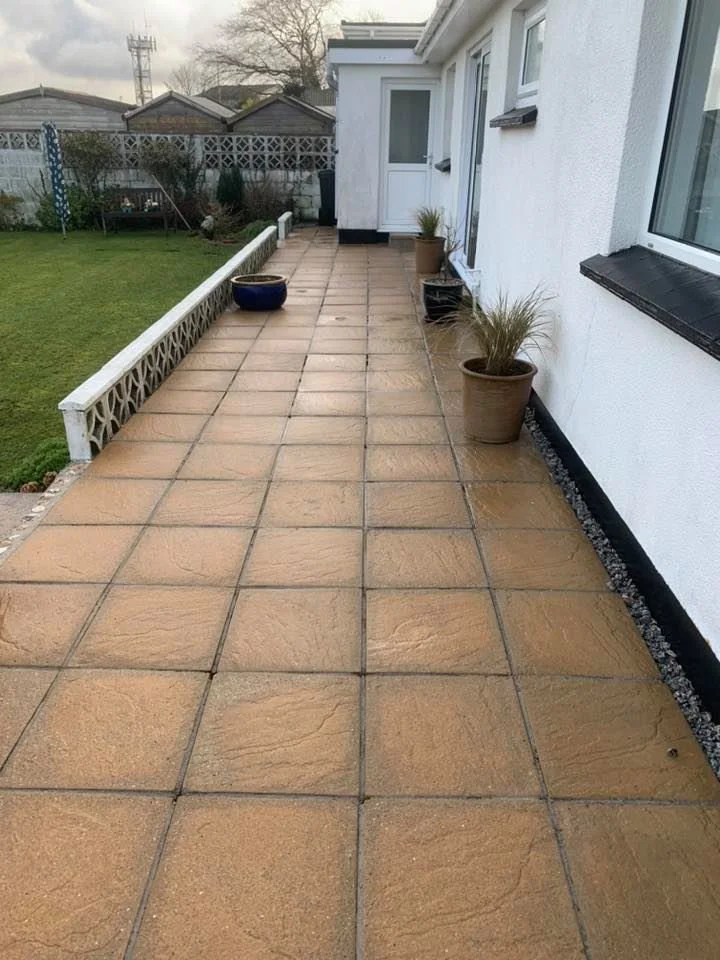 An outdoor patio with large tan square tiles, two potted plants along the white exterior wall, and a backyard with green grass and a white lattice fence.