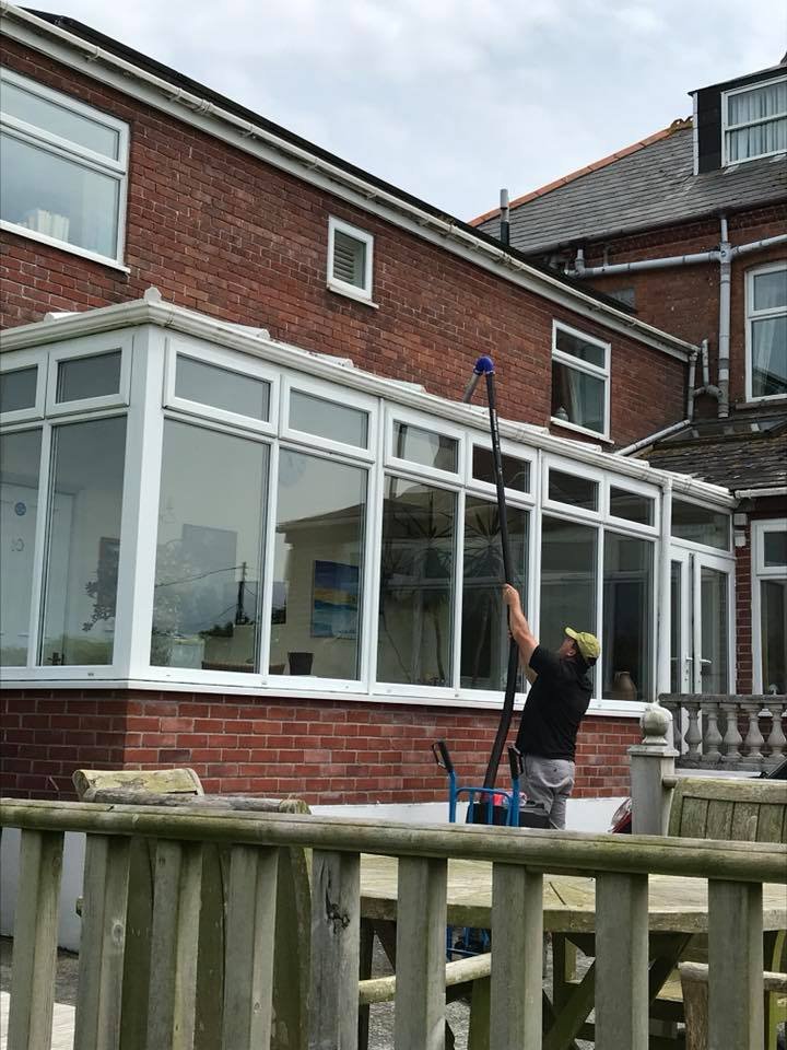 A man cleaning the outside of a conservatory attached to a brick house using a long pole with a cleaning tool at the end.