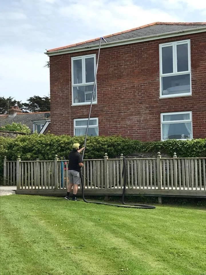 A person holding a long vacuum hose, cleaning or inspecting a second-story window on a brick house, with the hose extended diagonally upward to the window.