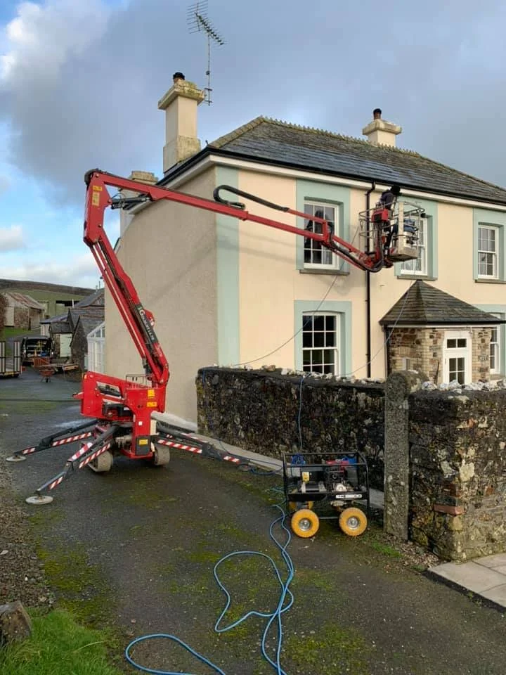 Worker in a lift cleaning the exterior window of a light-colored house with a gray roof, next to a stone wall.