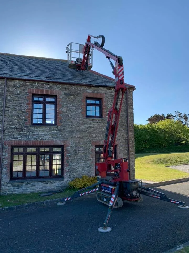 A cherry picker lift extended upward next to a brick house with stone walls and multiple windows. The lift is on a paved surface with stabilizing legs deployed.
