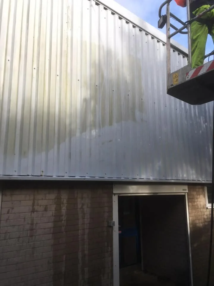 Construction worker on lift installing or repairing metal siding on the upper part of a building, with a brick wall and open door below.
