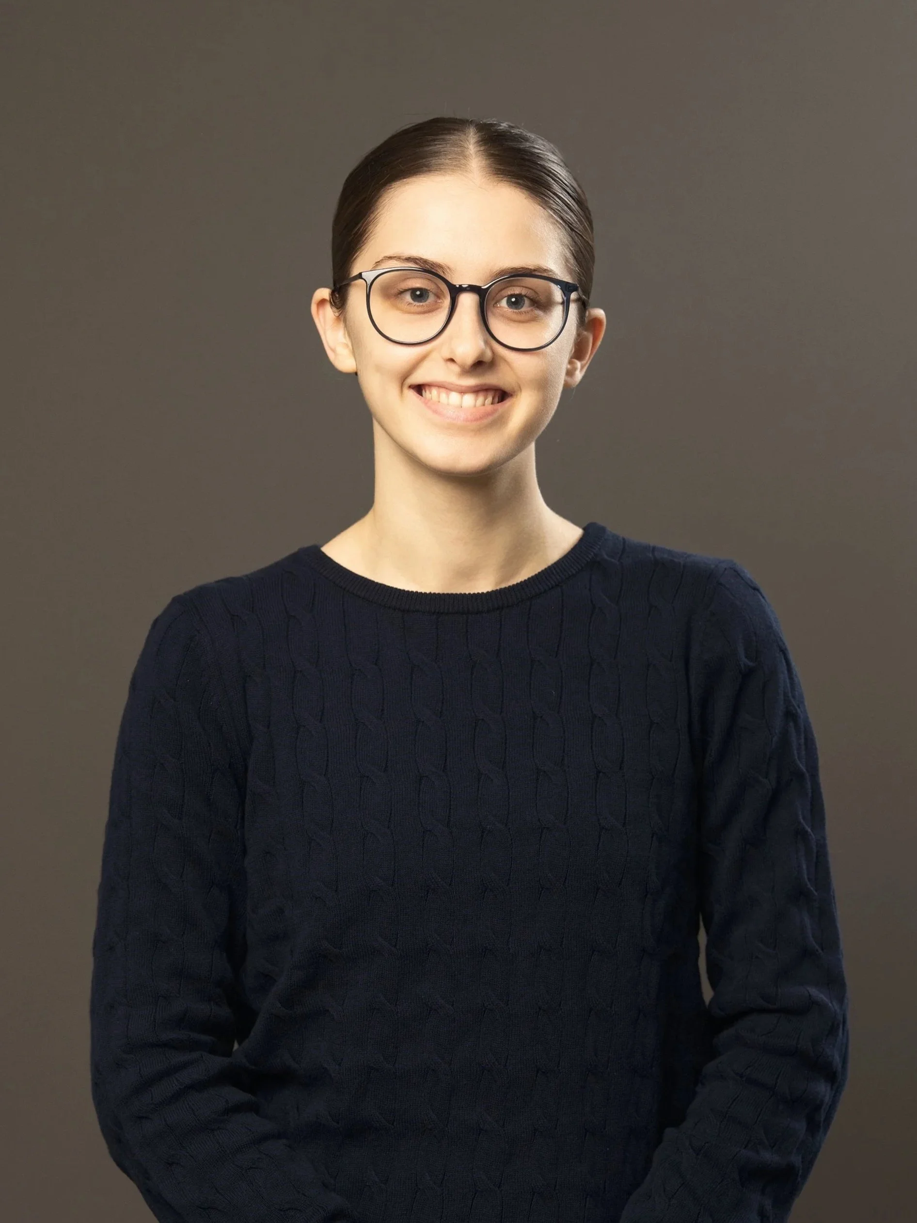 Young woman with glasses smiling, wearing a dark sweater, against a plain background.