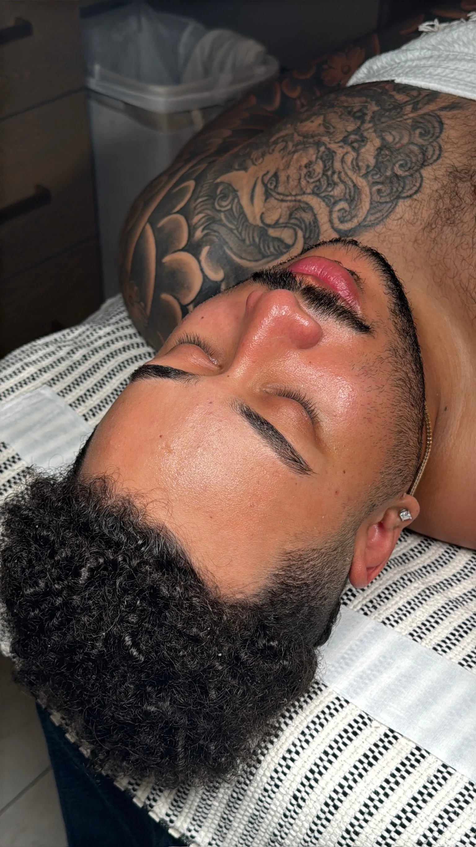 Close-up of a man with a fresh haircut, lying face up on a striped barber cape, with a large tattoo on his chest visible in the background.