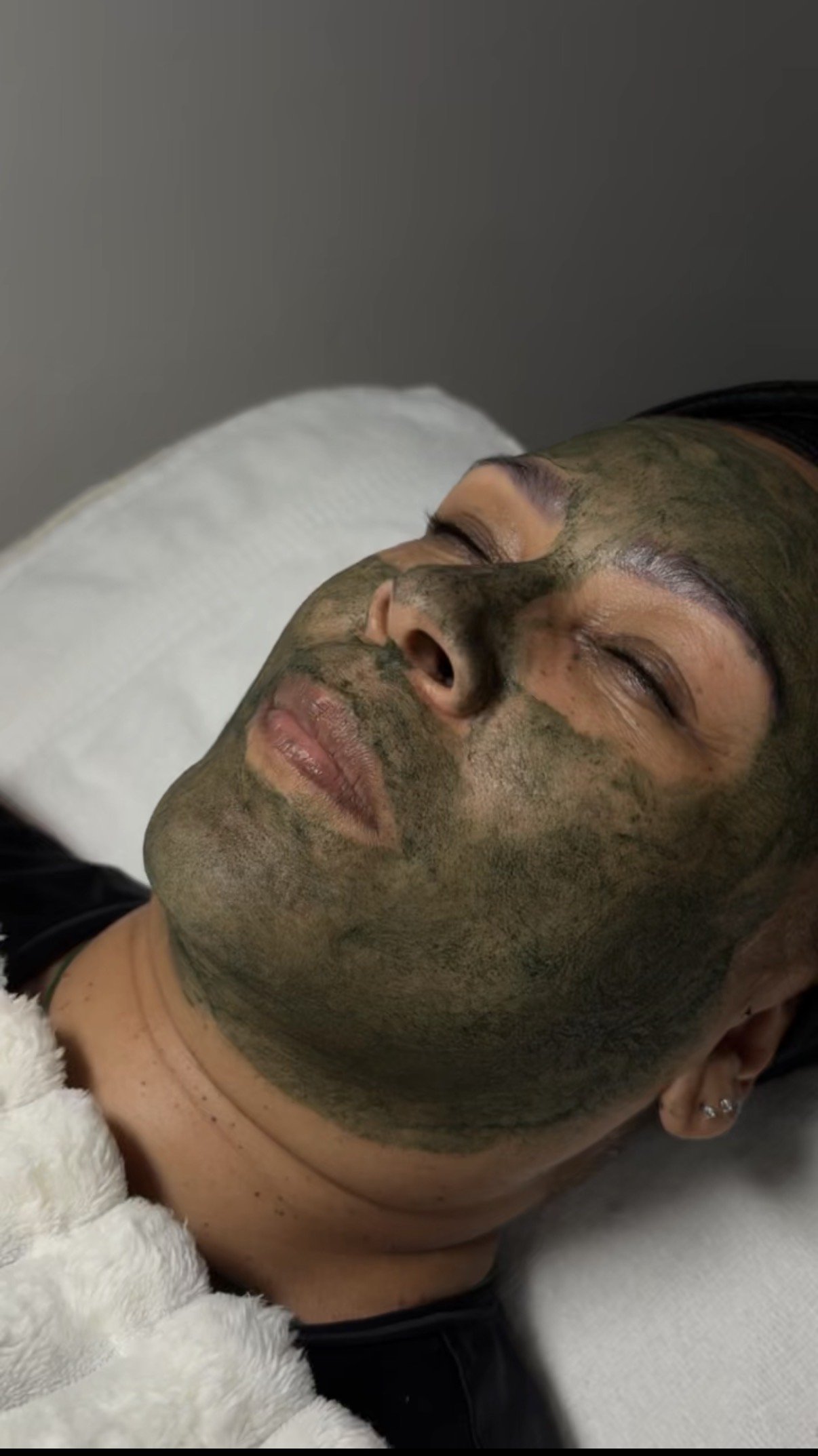 A person lying down with a green facial mask applied evenly across their face, eyes closed, appearing relaxed.