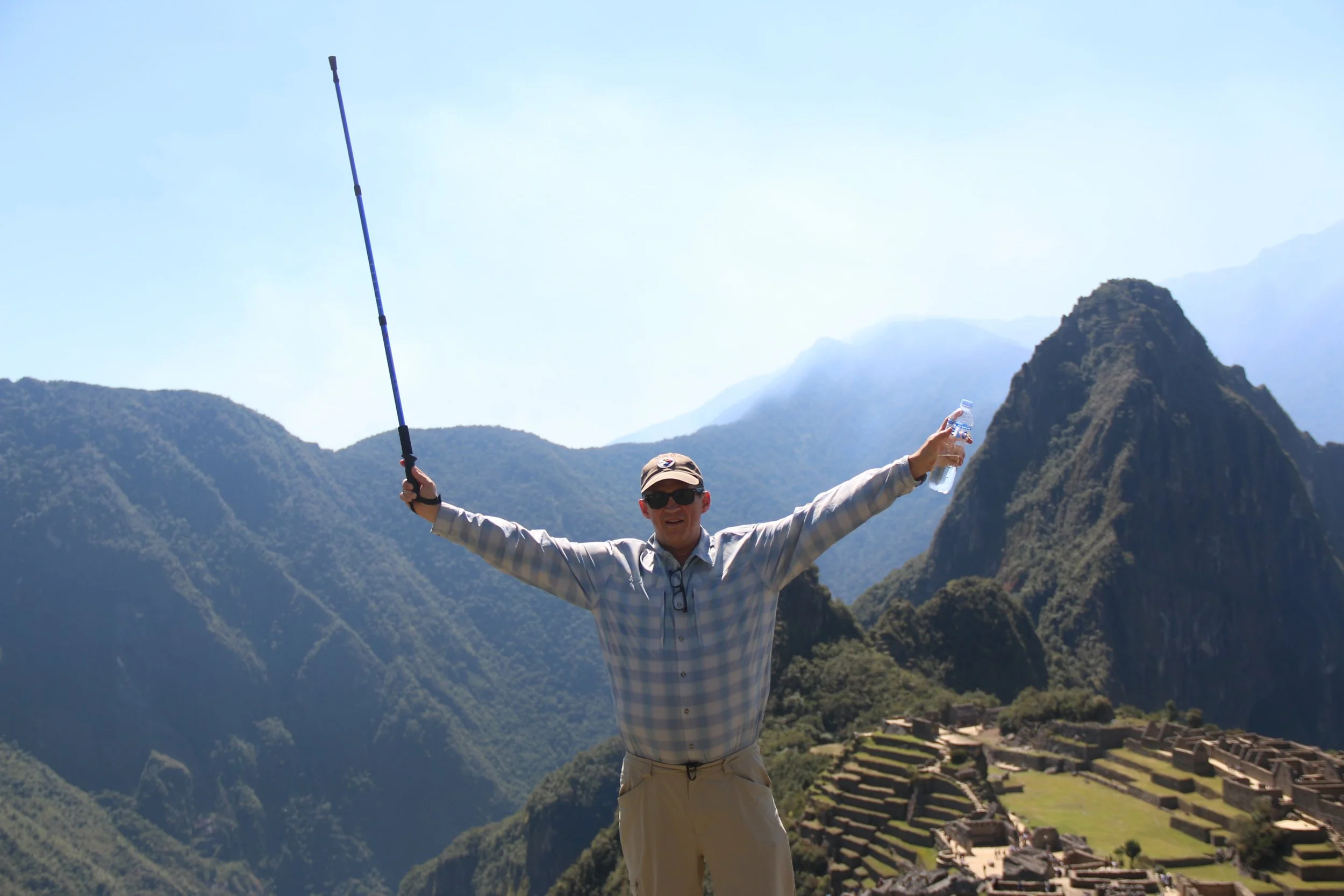 A man in sunglasses, a plaid shirt, and khaki pants standing with arms outstretched, holding a water bottle in one hand and a trekking pole in the other, in front of Machu Picchu and mountains.