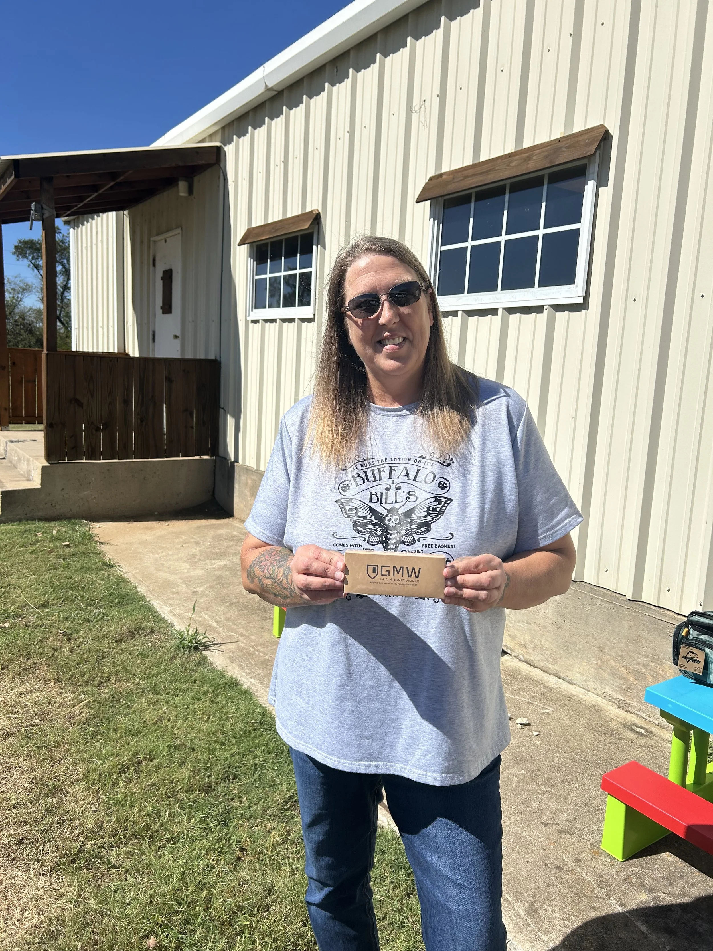 A woman with long hair wearing sunglasses, a gray Buffalo Bill's t-shirt, and jeans is standing outside in front of a light-colored building with windows. She is holding a cardboard box with the GMW logo. A colorful bench is visible on the right side of the image.